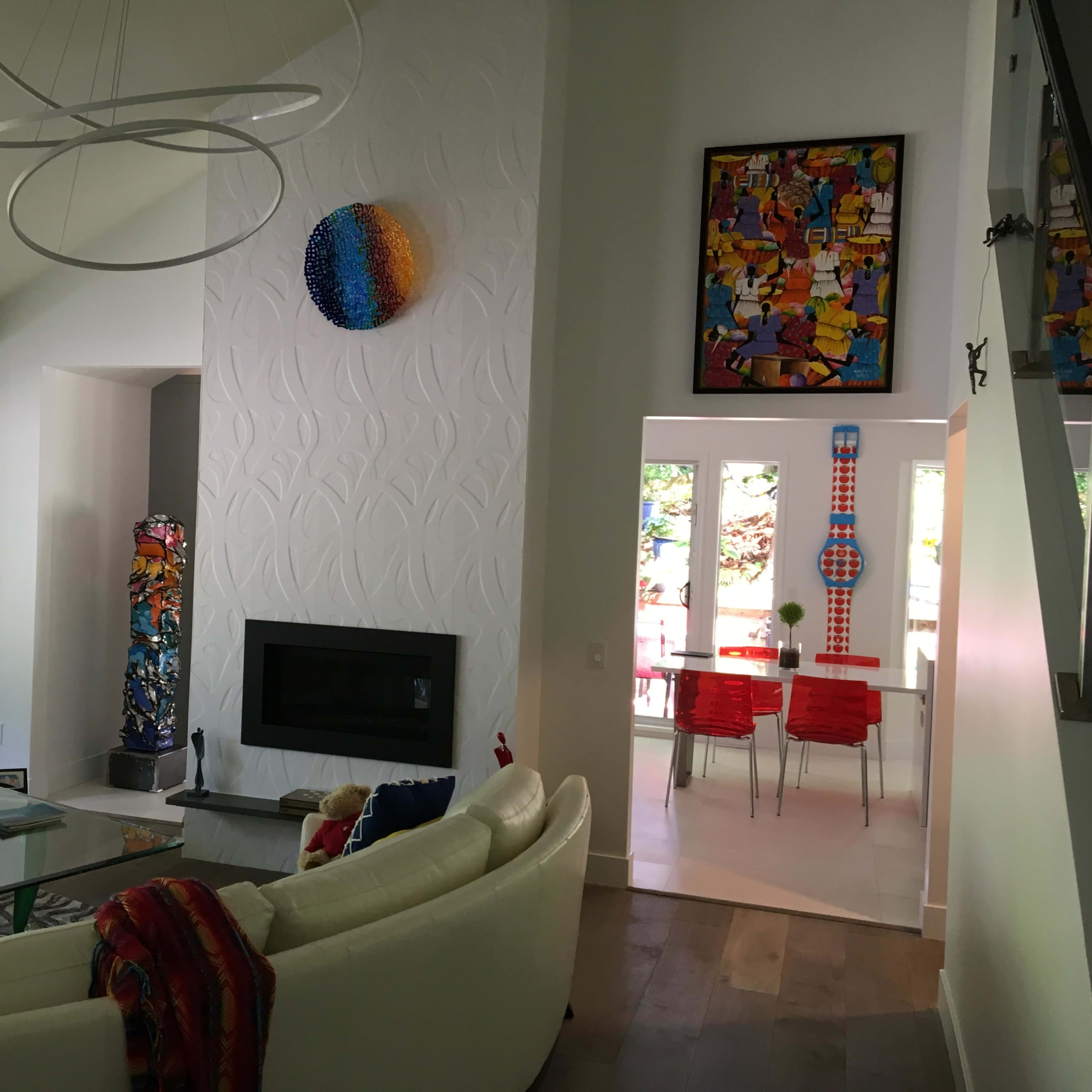 A bright, modern living room with a circular white sofa, a glass coffee table, a fireplace, and colorful artwork on the walls, leading into a dining area with red chairs.