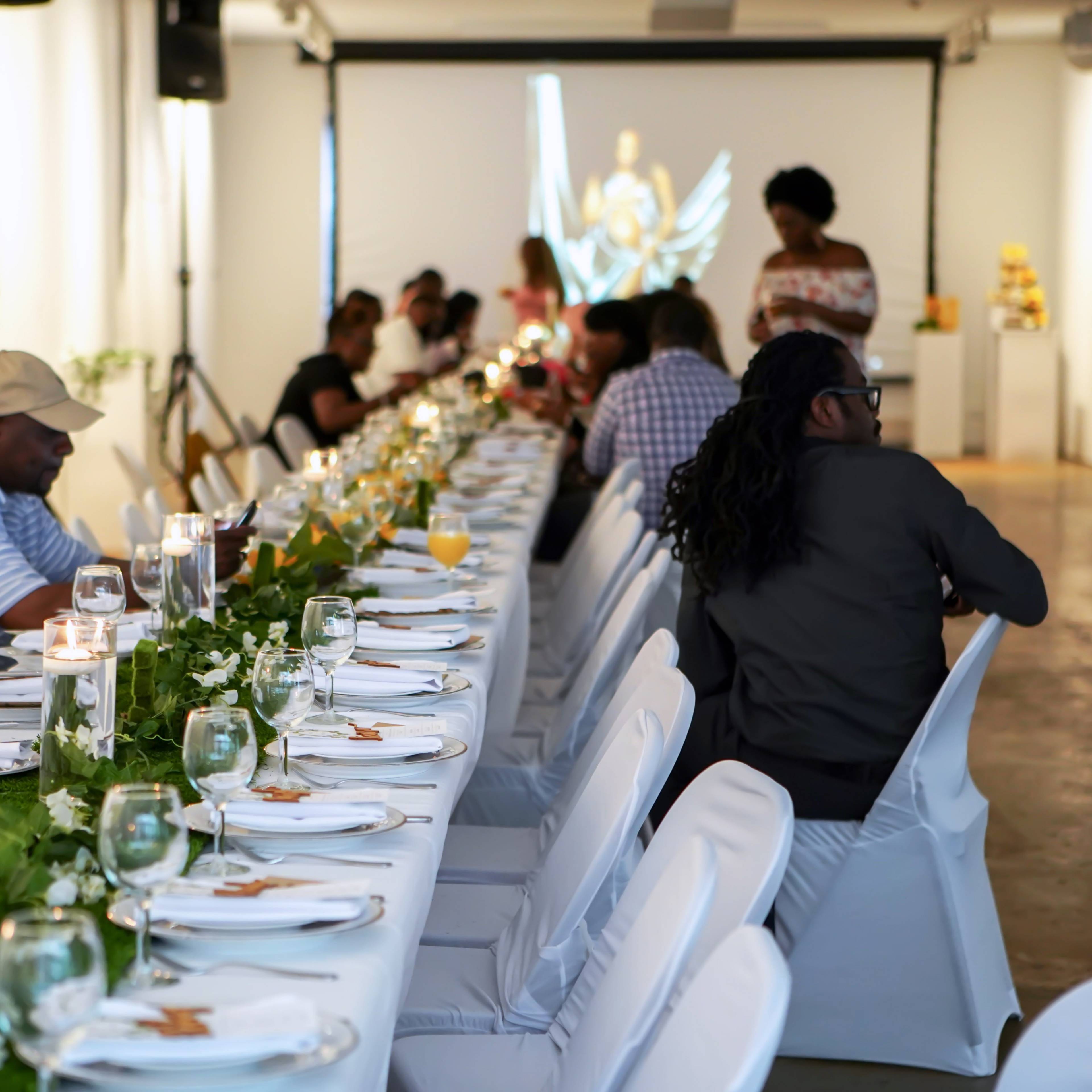 A long dining table decorated with greenery and candles is set up in a well-lit room where guests are seated, engaging in conversation.