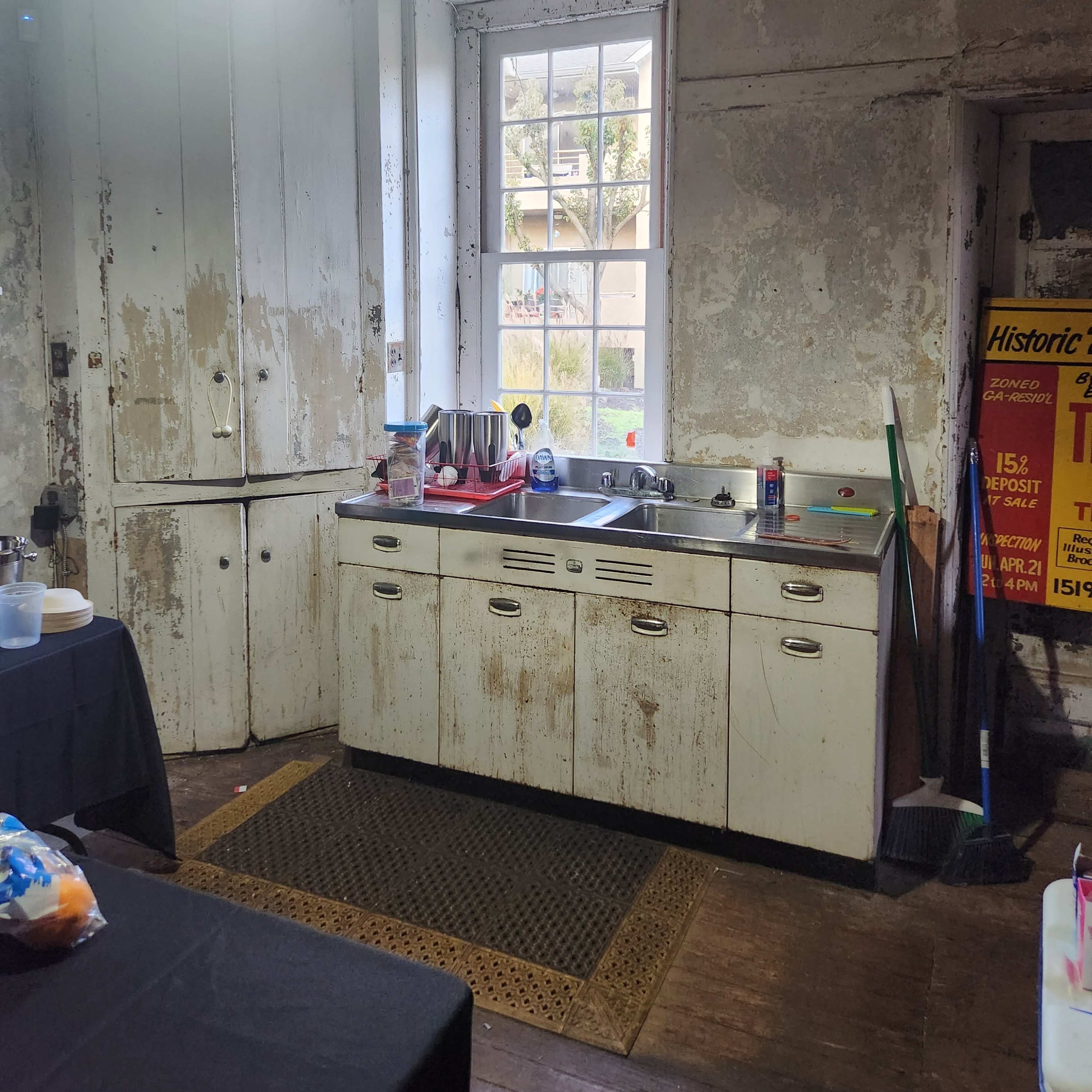 A weathered kitchen with a vintage sink, a window allowing natural light, and various cleaning tools nearby on the floor.