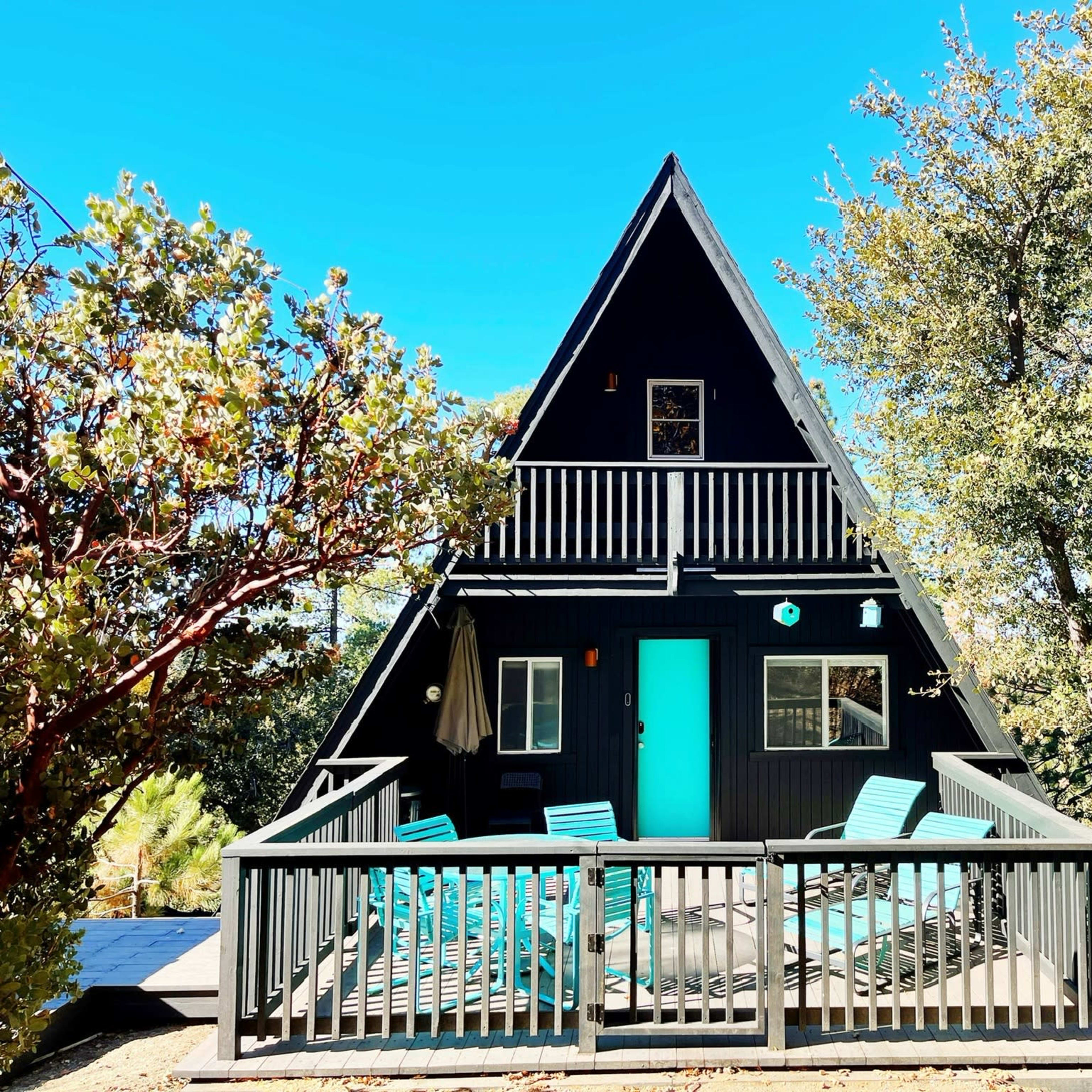 A black A-frame house with a turquoise door and a surrounding deck is nestled among trees in a clear blue sky.