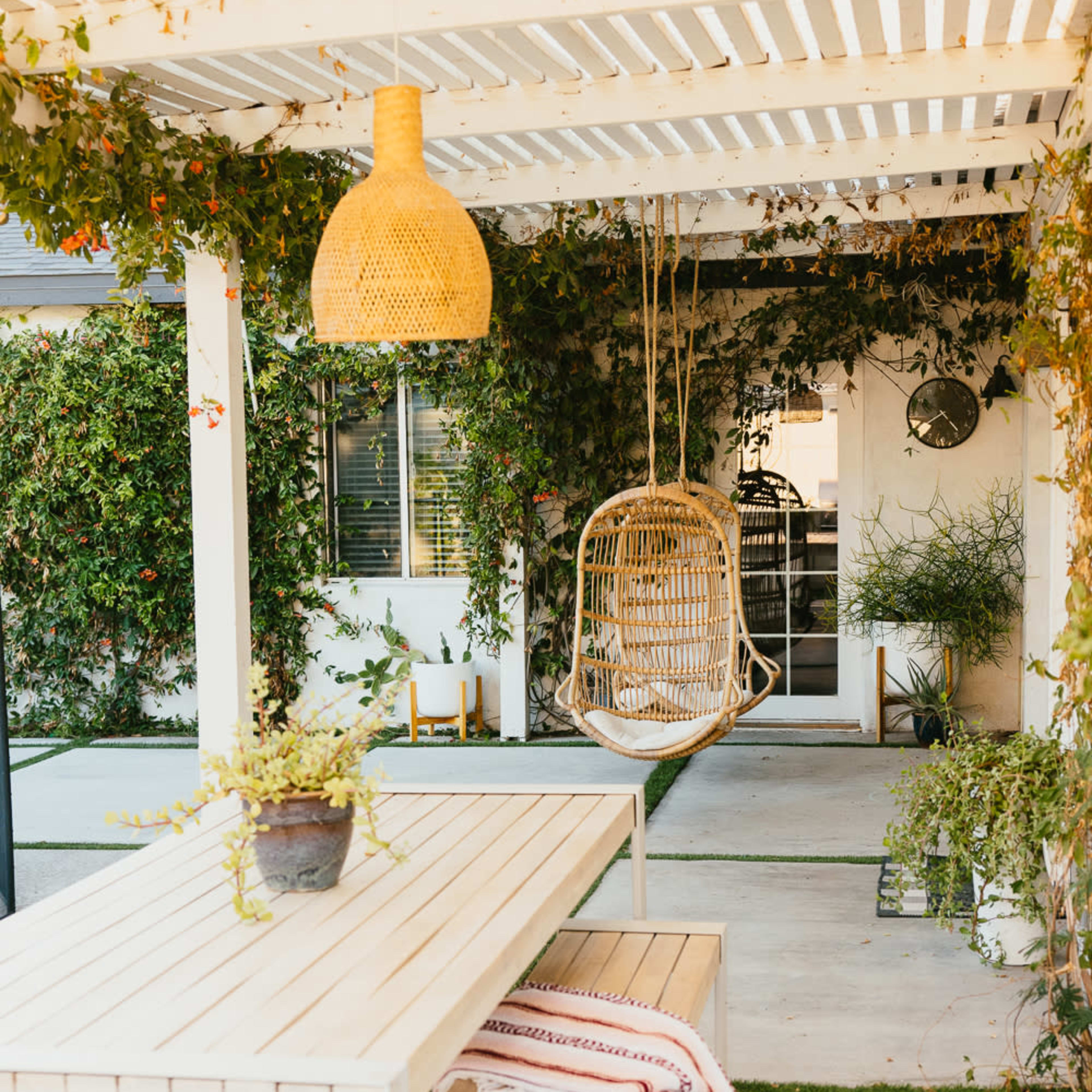 A covered outdoor patio features a wooden table and benches, a hanging wicker chair, and lush greenery along the walls.