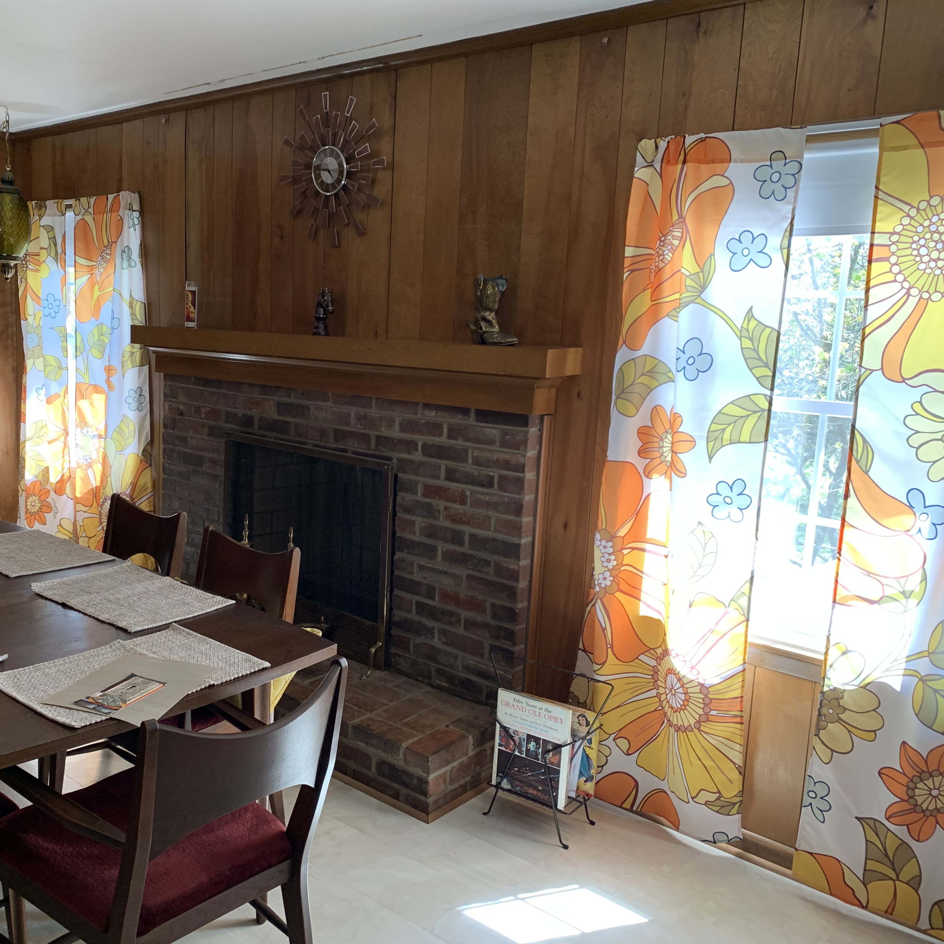 A dining room with a wooden table surrounded by chairs, a brick fireplace, and floral-patterned curtains on two windows.