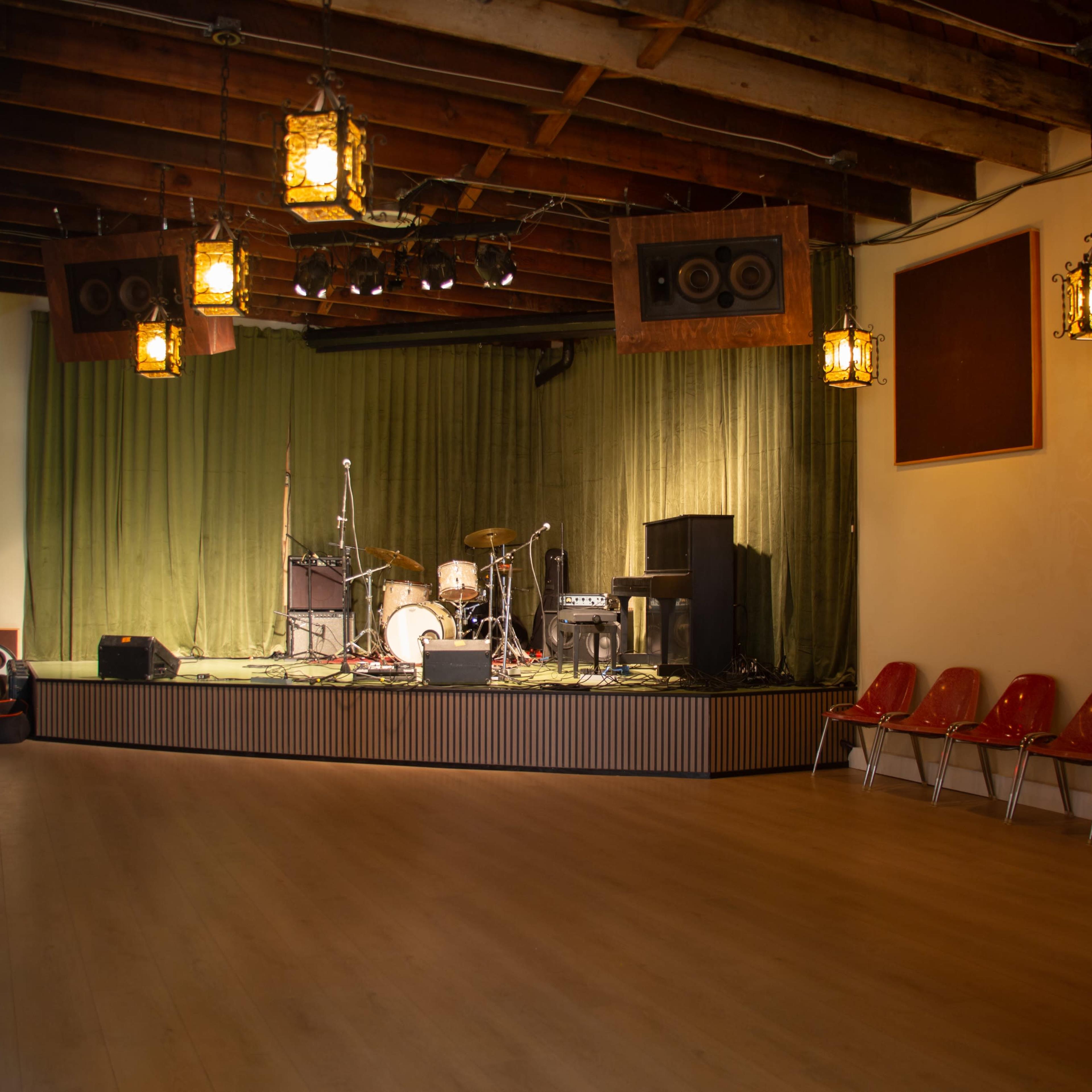 A stage set up for a performance, featuring musical instruments, a green backdrop, and several empty red chairs arranged in front of it.
