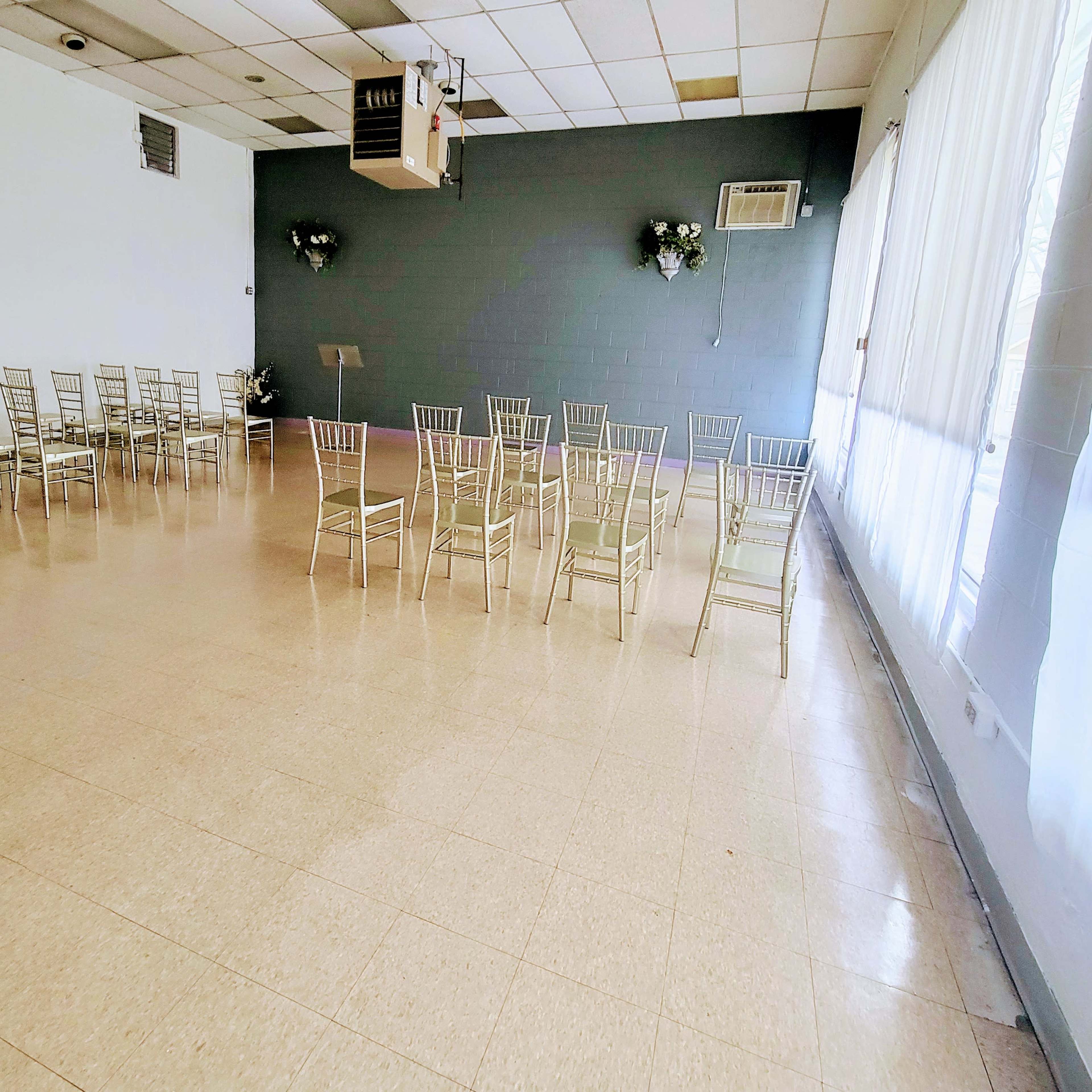 A spacious, empty room with rows of metal chairs arranged facing a blank wall, large windows allowing natural light to enter.