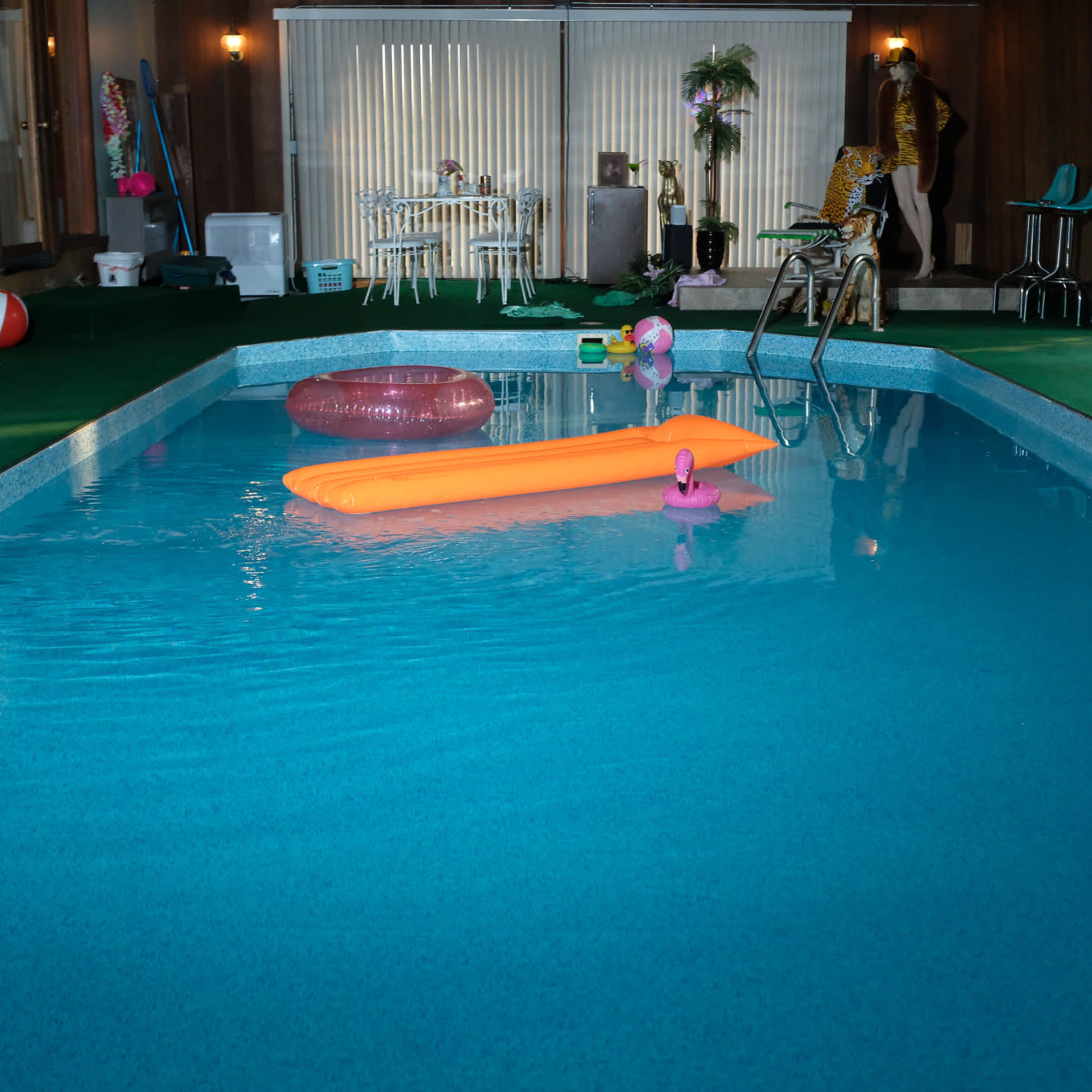An indoor pool surrounded by green carpet, with a pink flamingo float and an orange float in the water, and a decorative area featuring furniture and plants in the background.