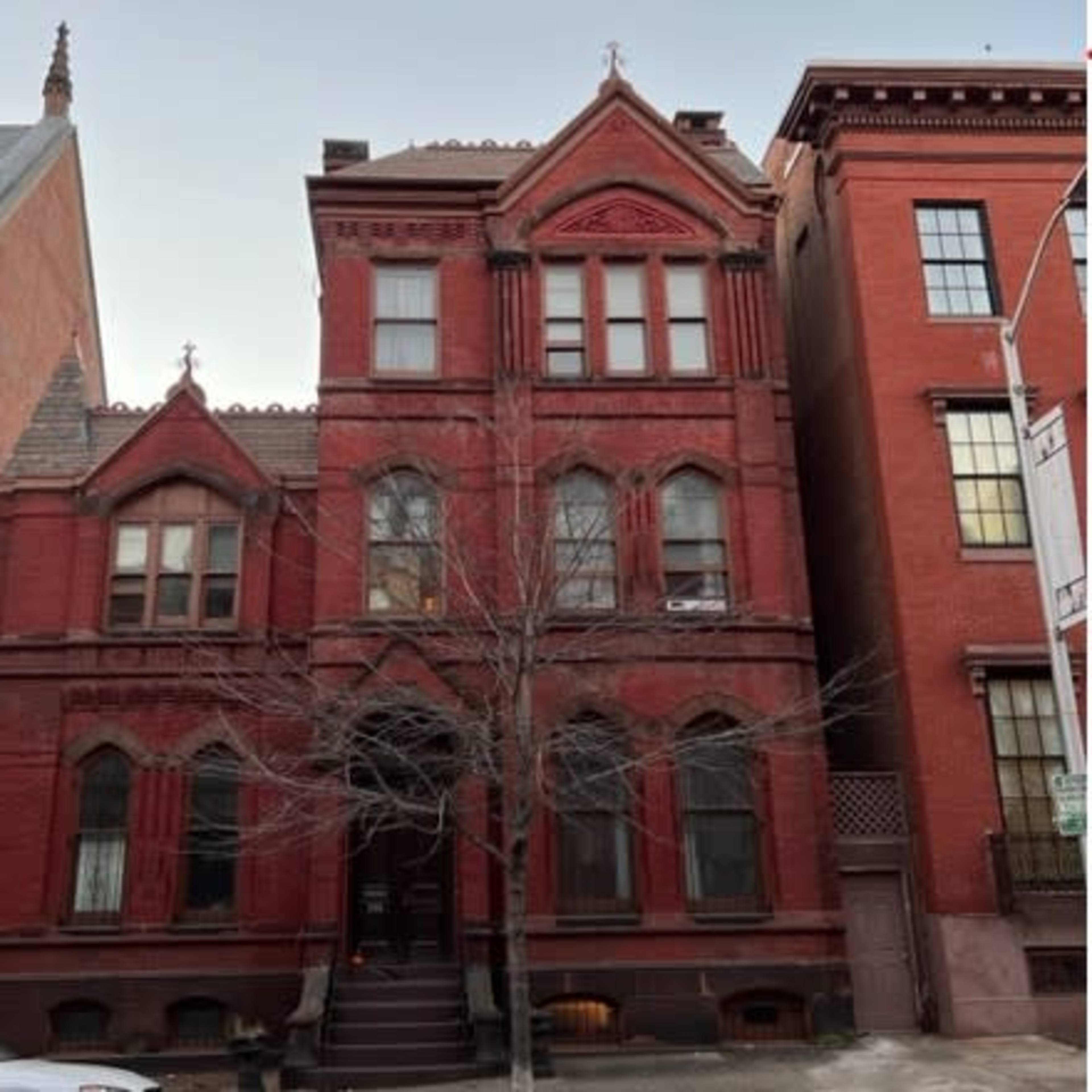 A three-story red brick Victorian-style house with decorative elements, flanked by two taller brick buildings.