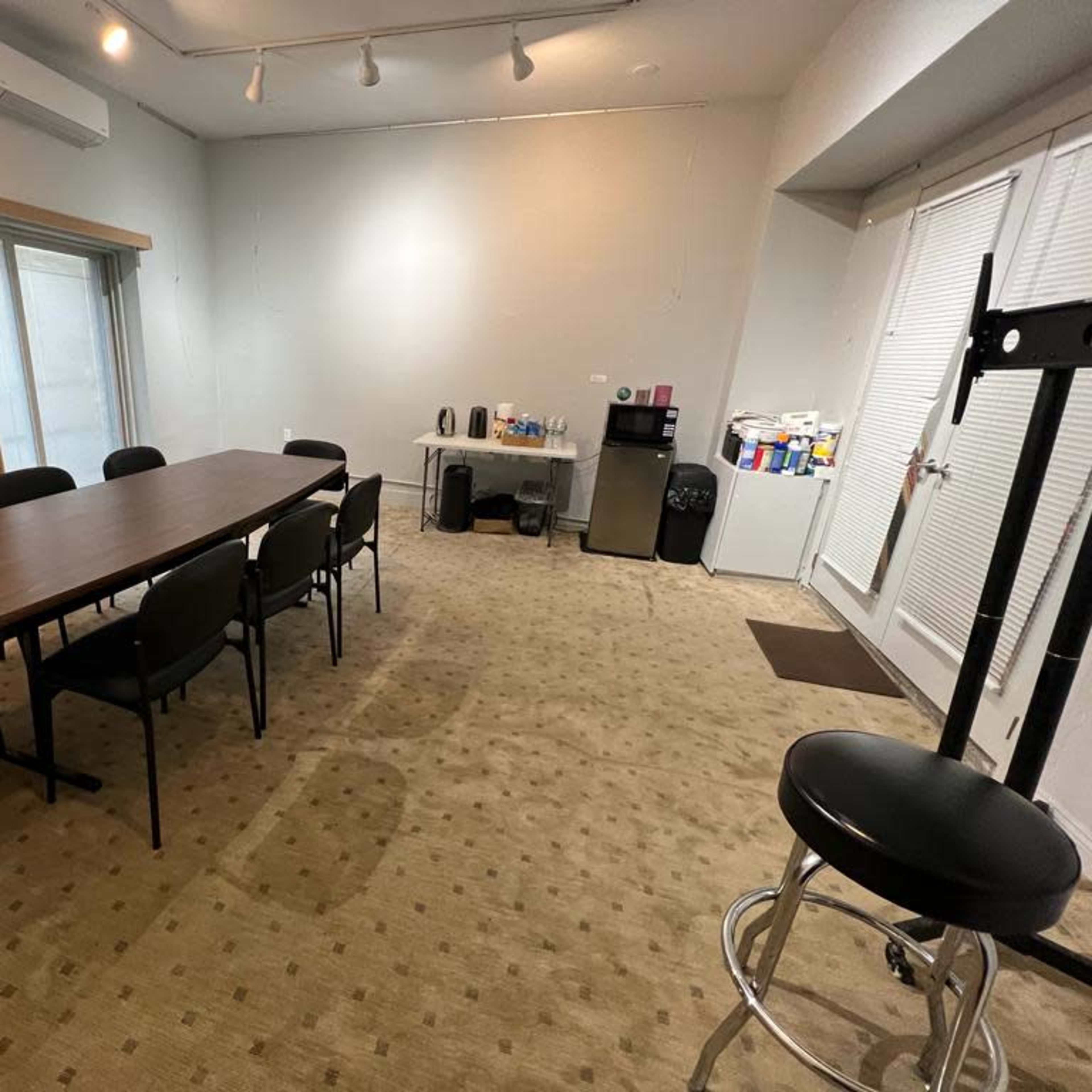 A conference room features a long table surrounded by chairs, with a kitchenette area and a large window in the background.