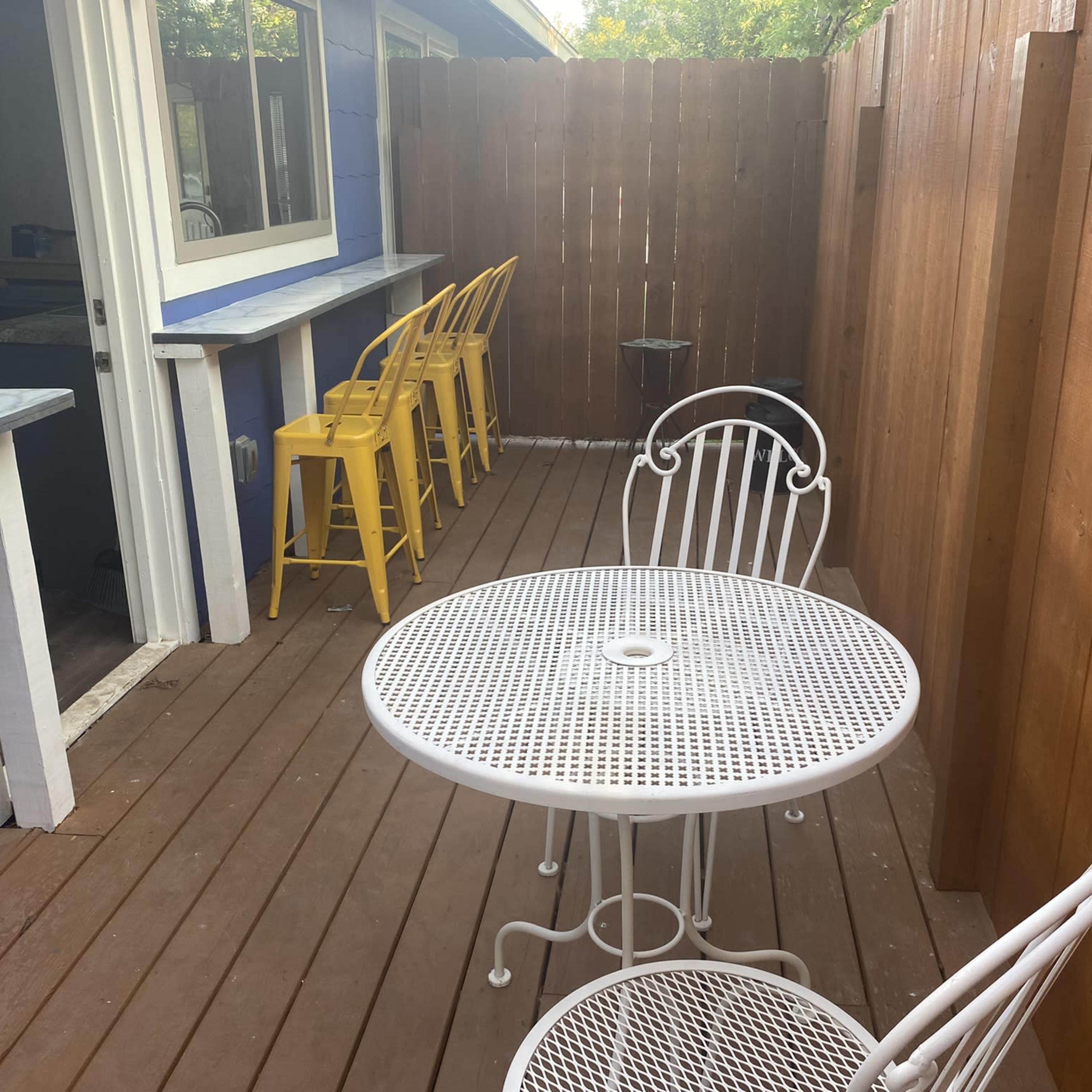 A small outdoor patio features a round white metal table and chairs, along with yellow bar stools against a wooden fence.