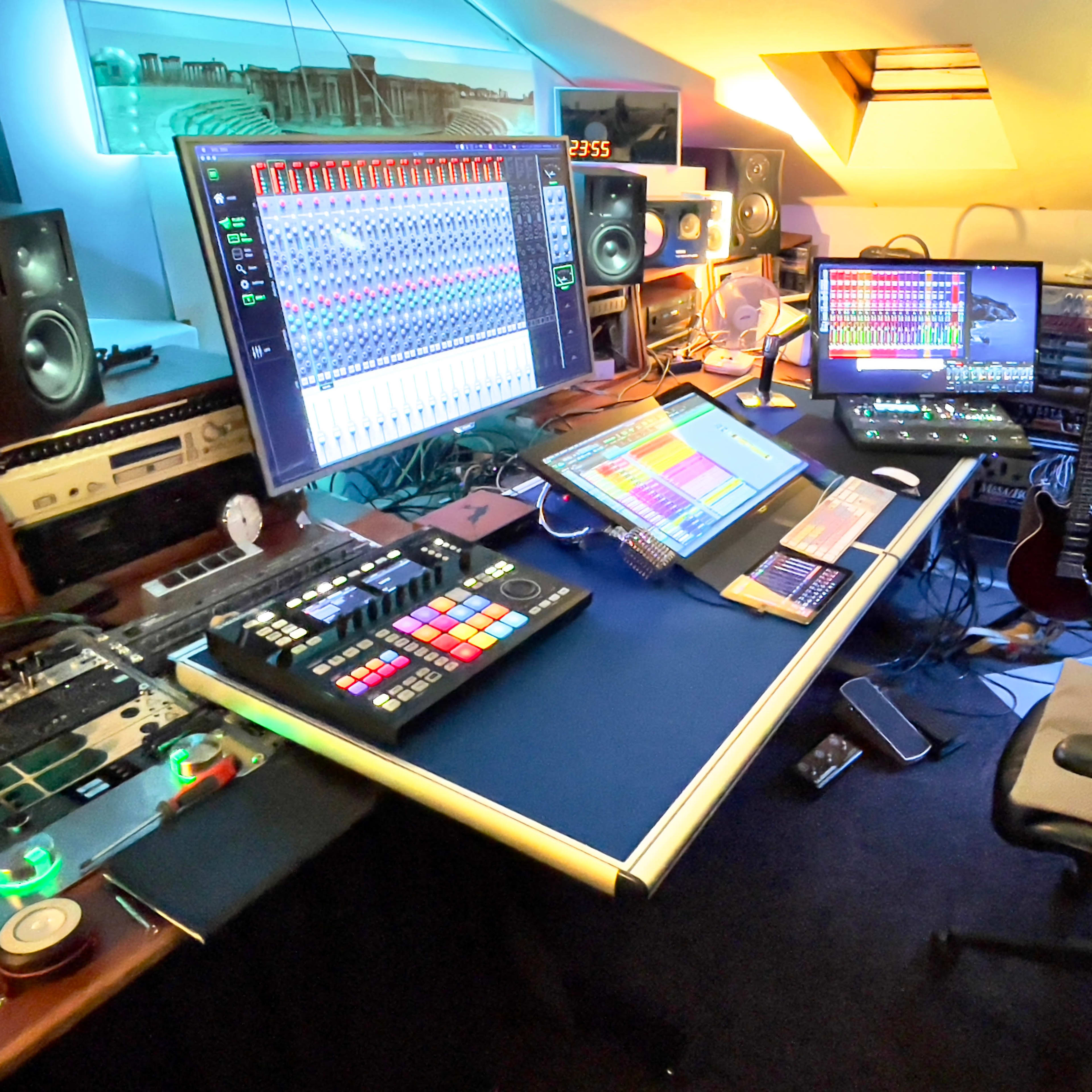A modern home music studio with multiple screens, audio equipment, and musical instruments arranged on a desk.
