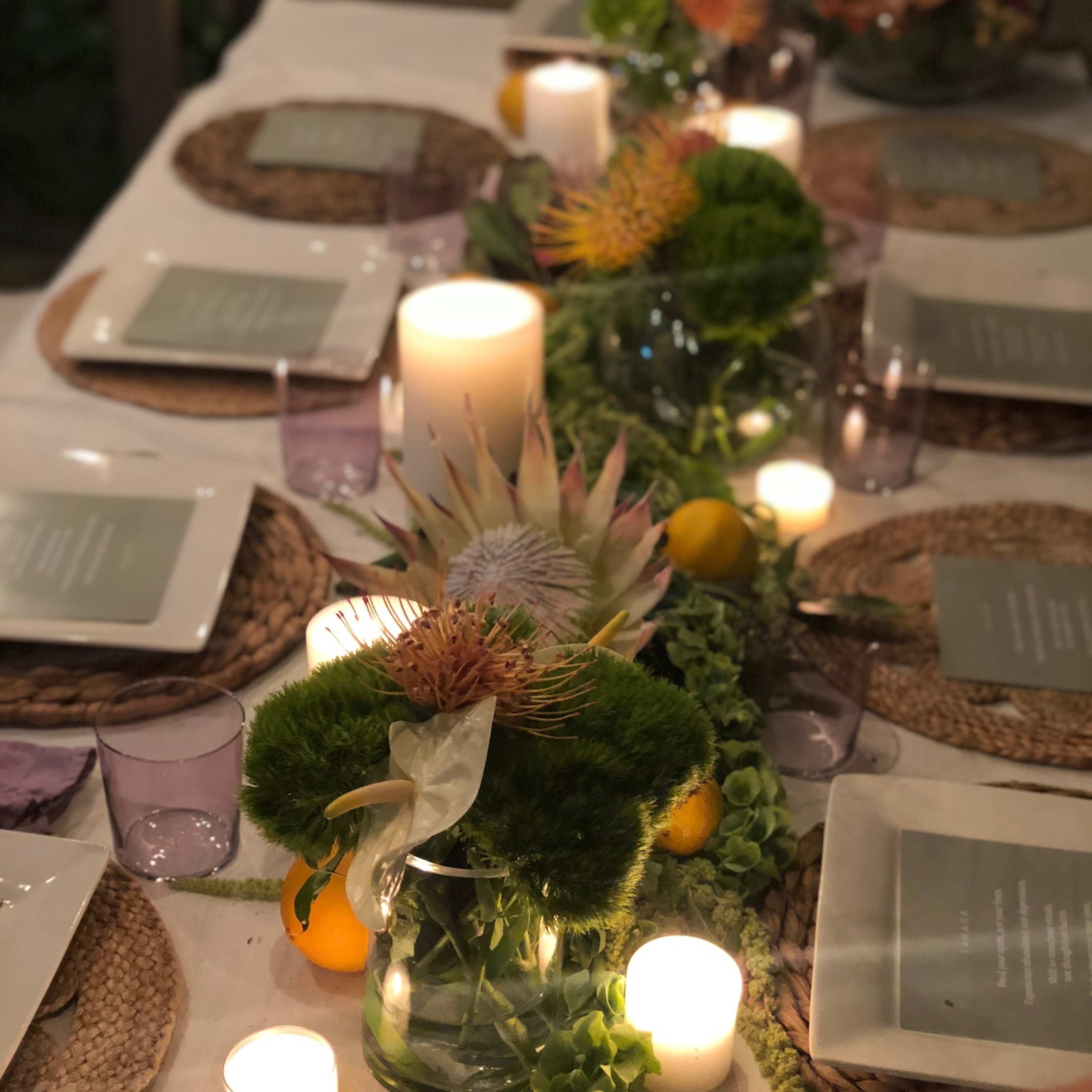 A dinner table is set with white plates, menus, candles, and floral arrangements, surrounded by woven placemats and garnished with greenery and fruits.