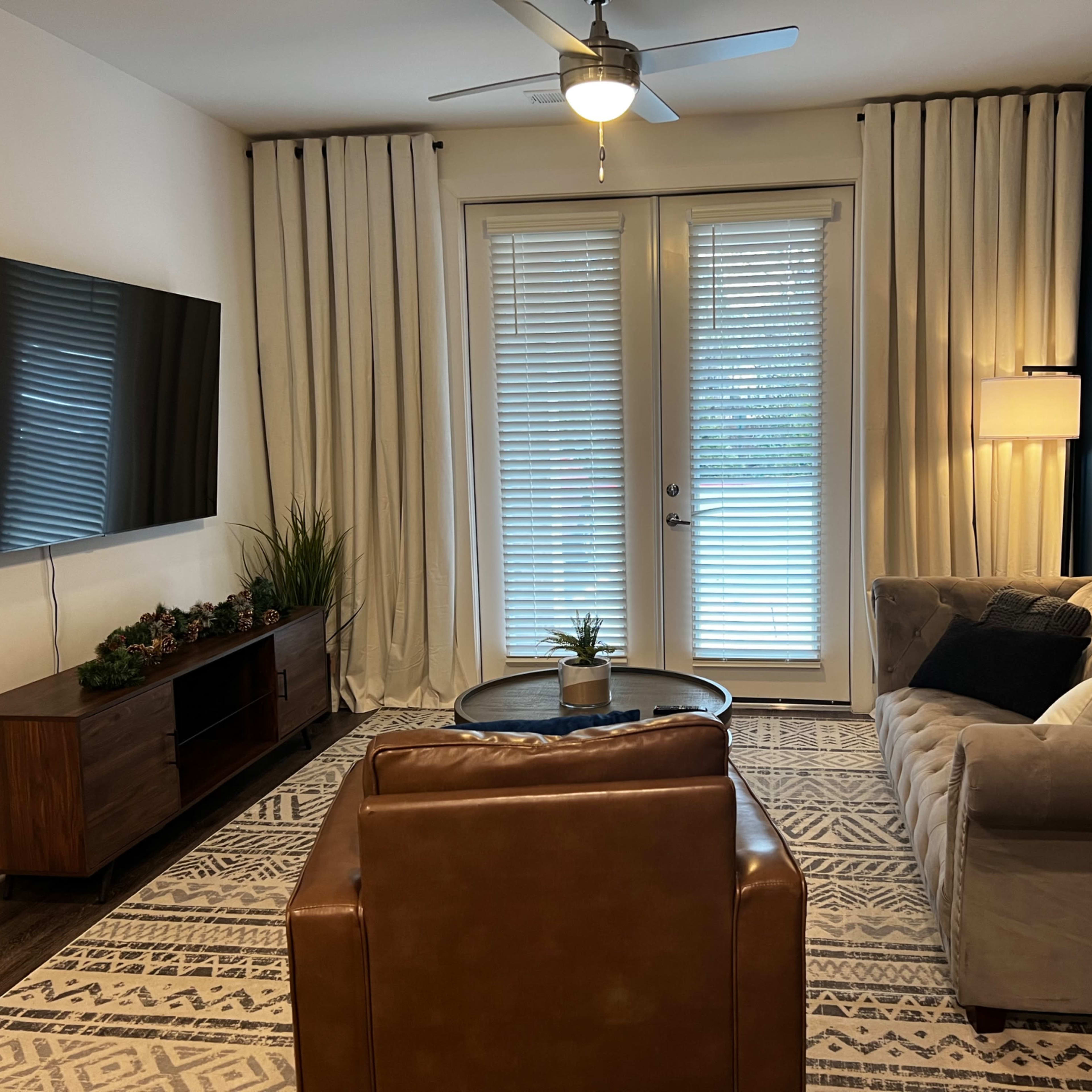 The living room features a television mounted on the wall, a beige sofa, a brown armchair, and large windows covered with sheer curtains.