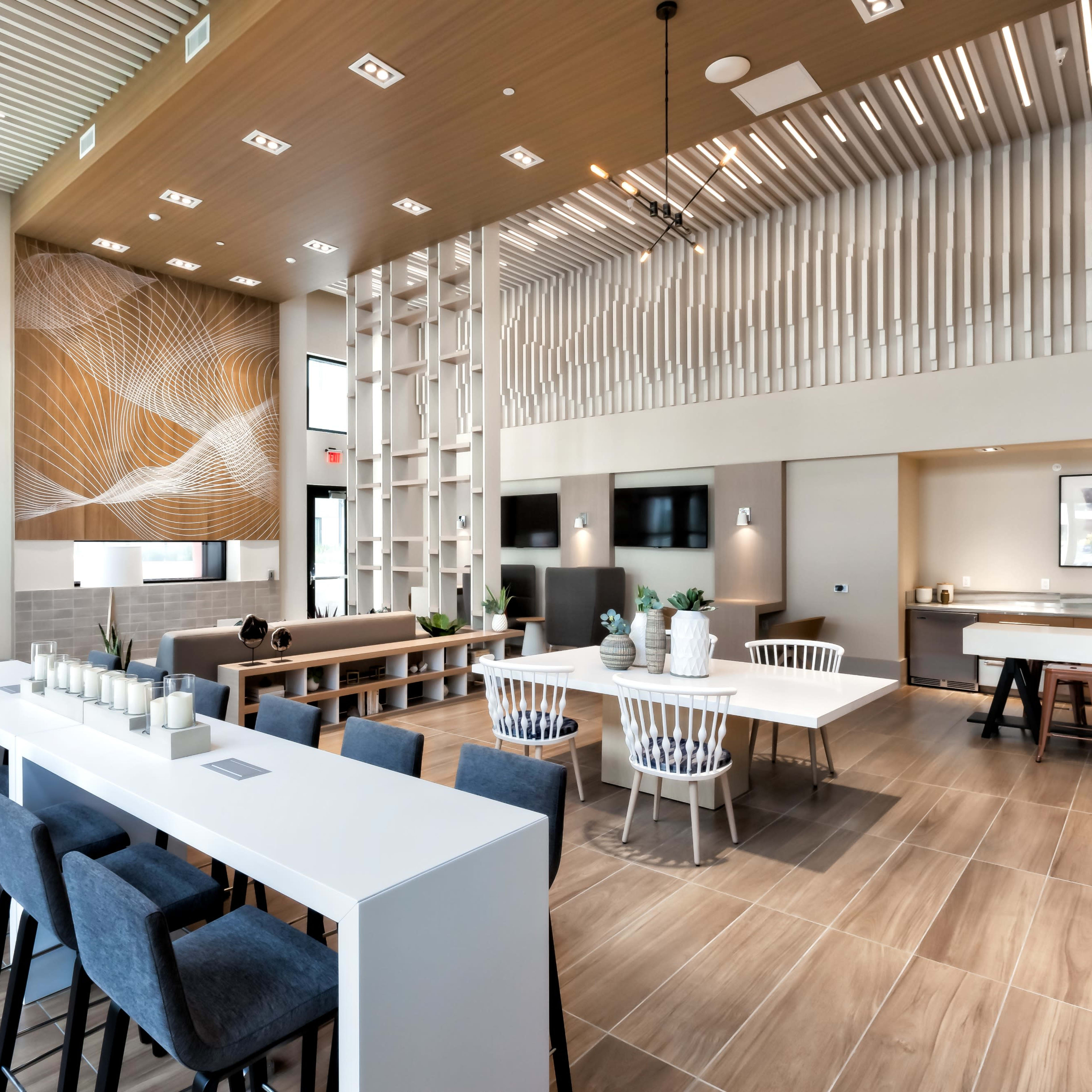 An interior communal lounge area with modern furniture, large windows, and a mix of seating arrangements around a long table and a bar-height counter.
