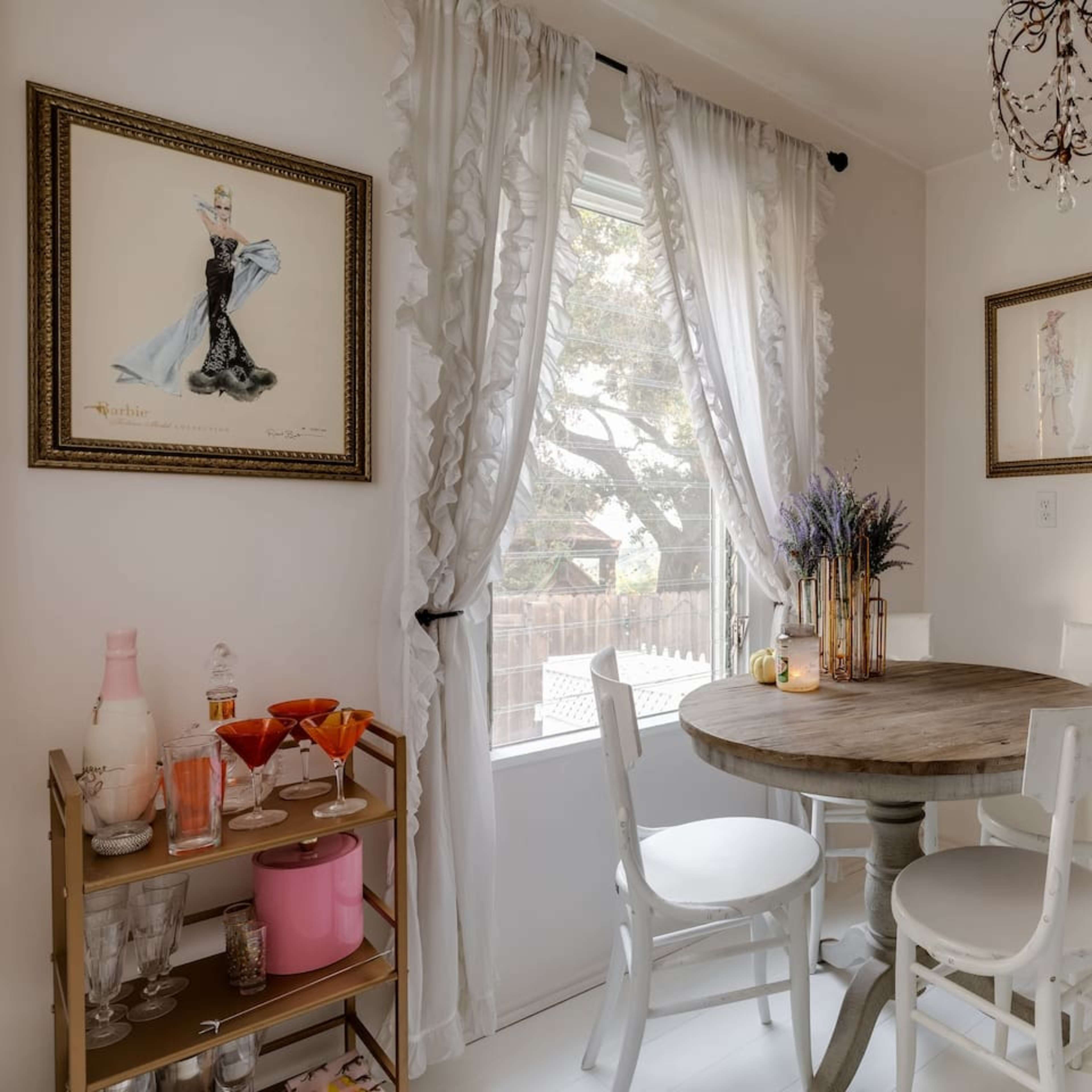 A small dining area features a round wooden table surrounded by white chairs, with window drapes and framed artwork on the walls.