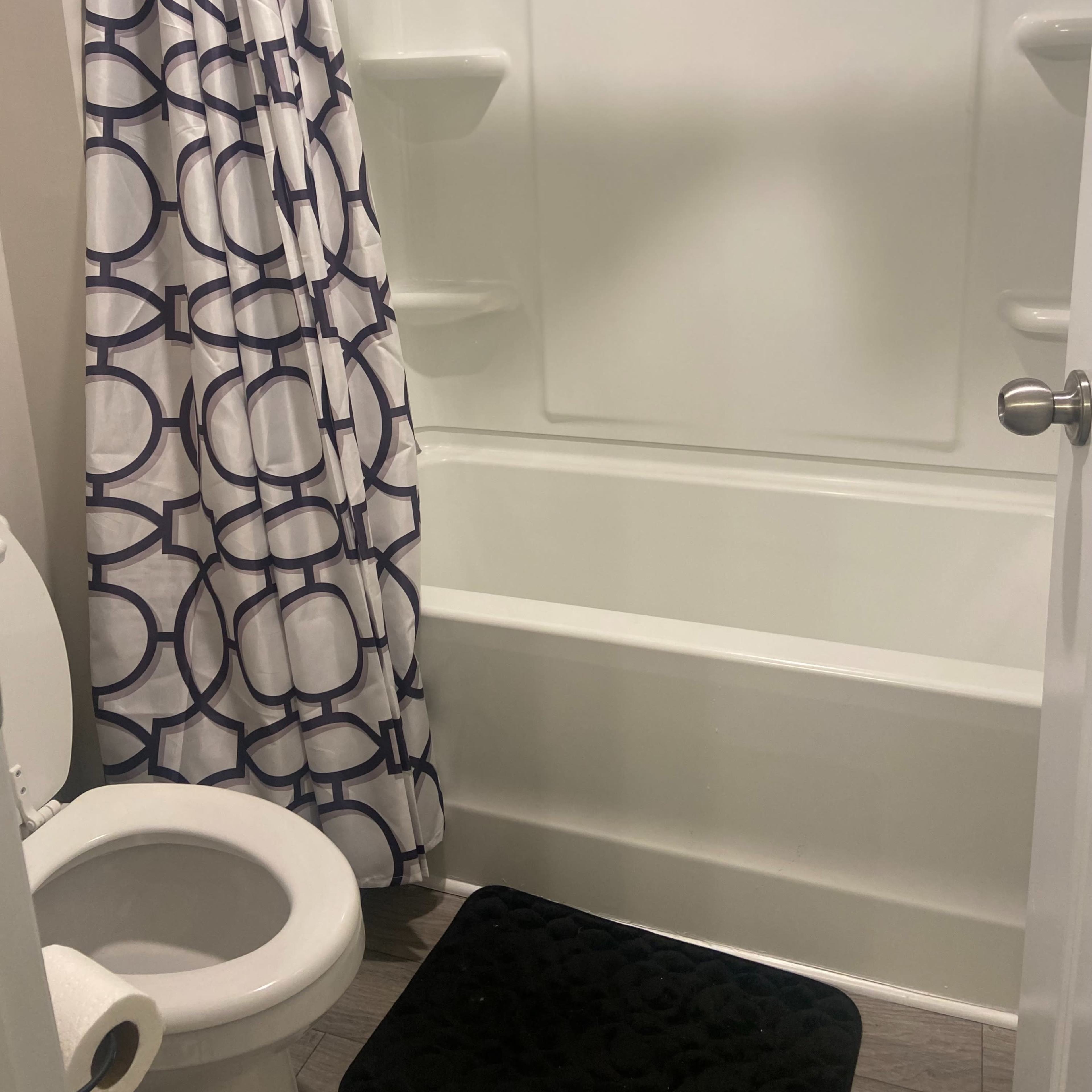 A bathroom with a toilet, a shower area with a patterned curtain, and a black mat on the floor.