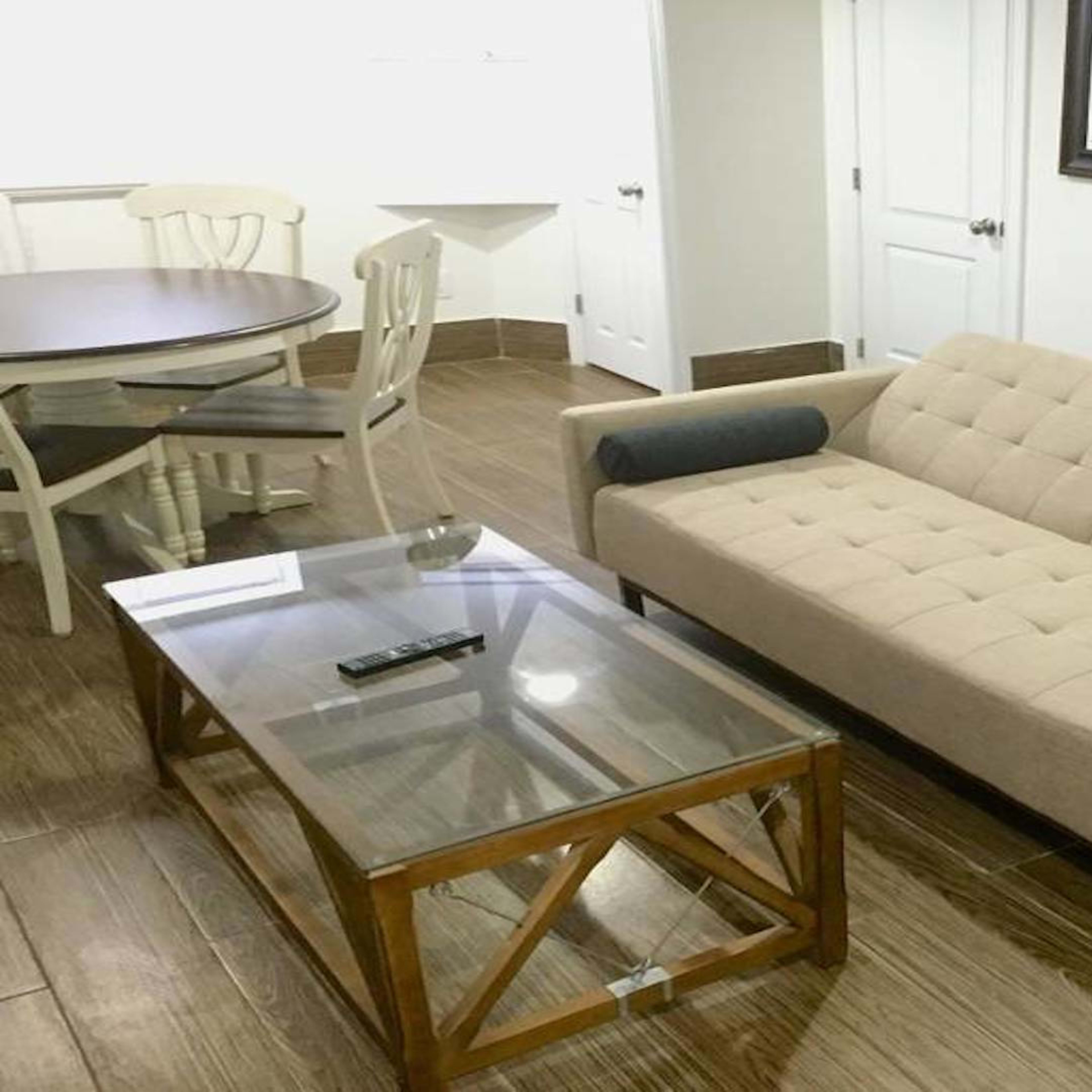 A light-colored sofa with dual arm cushions is placed in a room featuring a glass coffee table and a round dining table with white chairs.