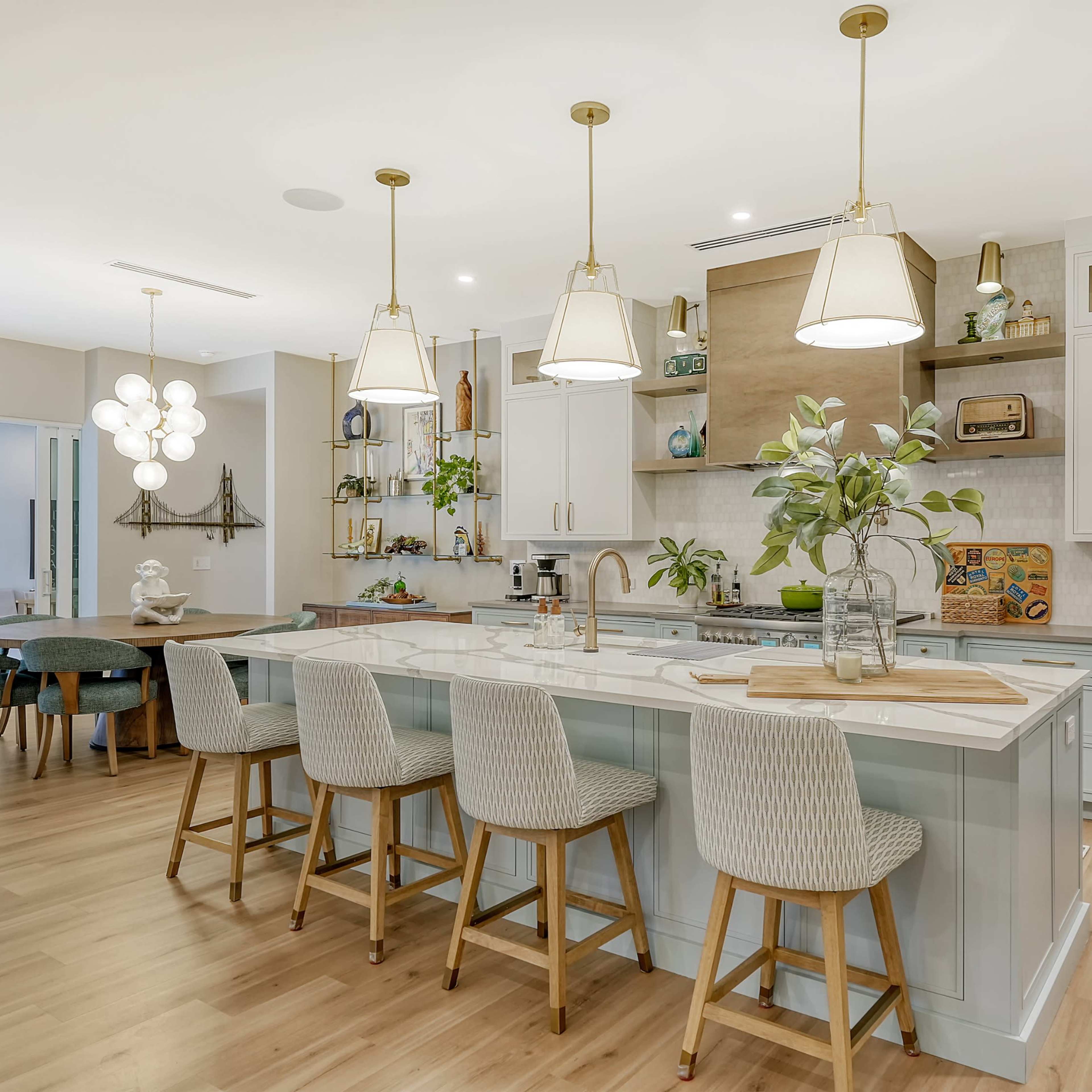 A modern kitchen features a large central island with bar stools, light wood flooring, and various plants and decor elements.