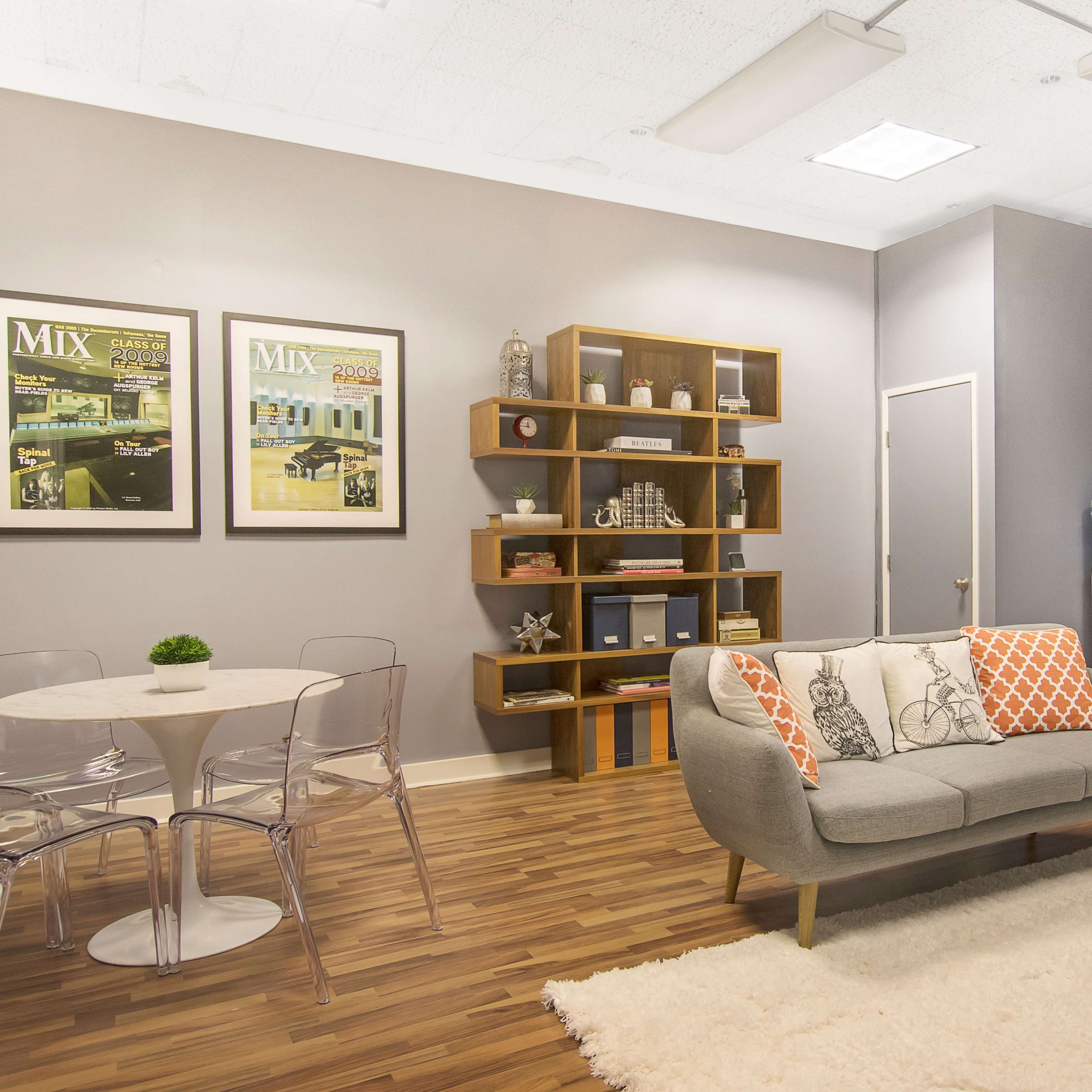 A modern lounge area with a circular dining table, transparent chairs, a gray sofa with orange and white pillows, a bookcase, and a kitchenette with a water dispenser and fridge.