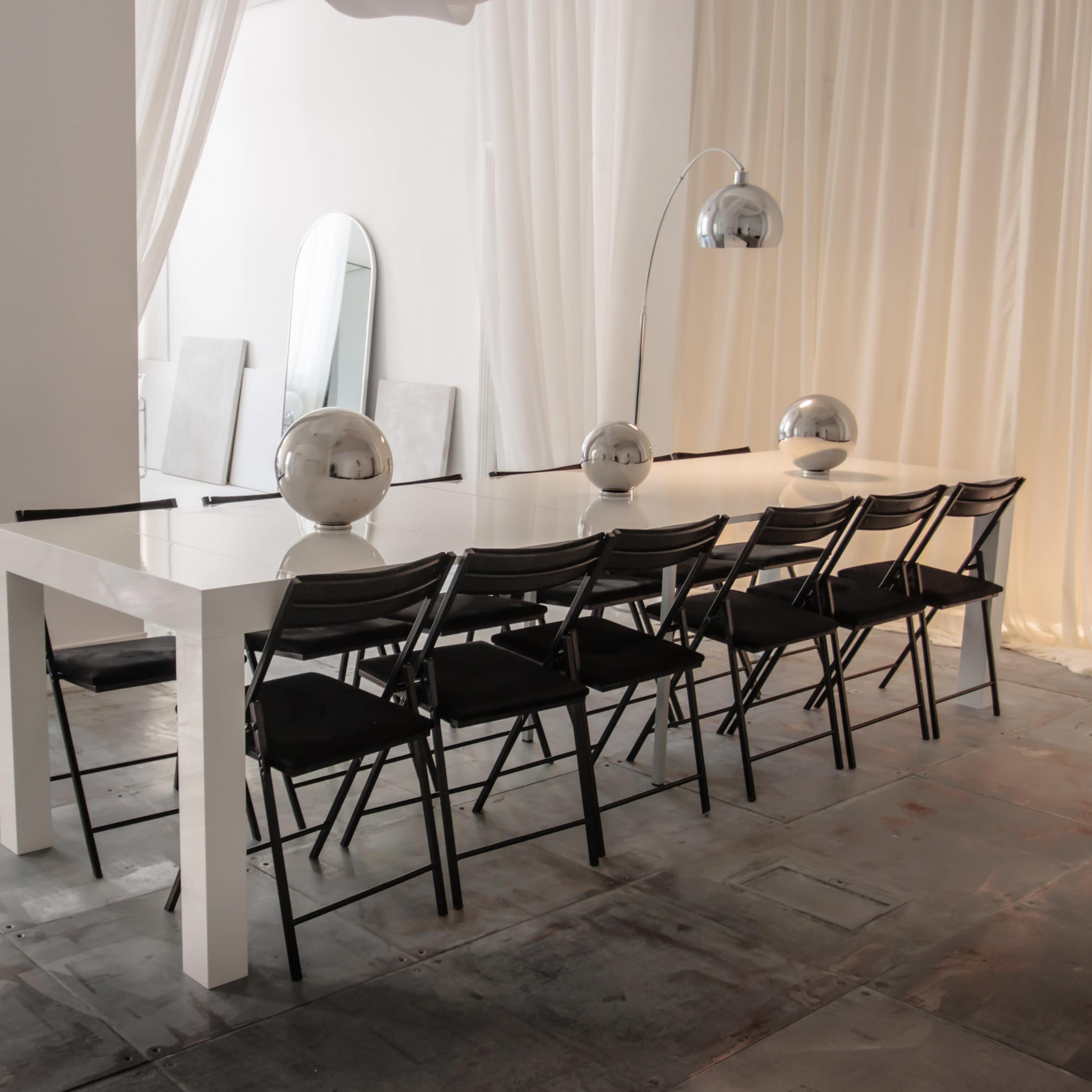 A long white dining table with black chairs is set in a brightly lit room adorned with mirrors and sheer curtains.