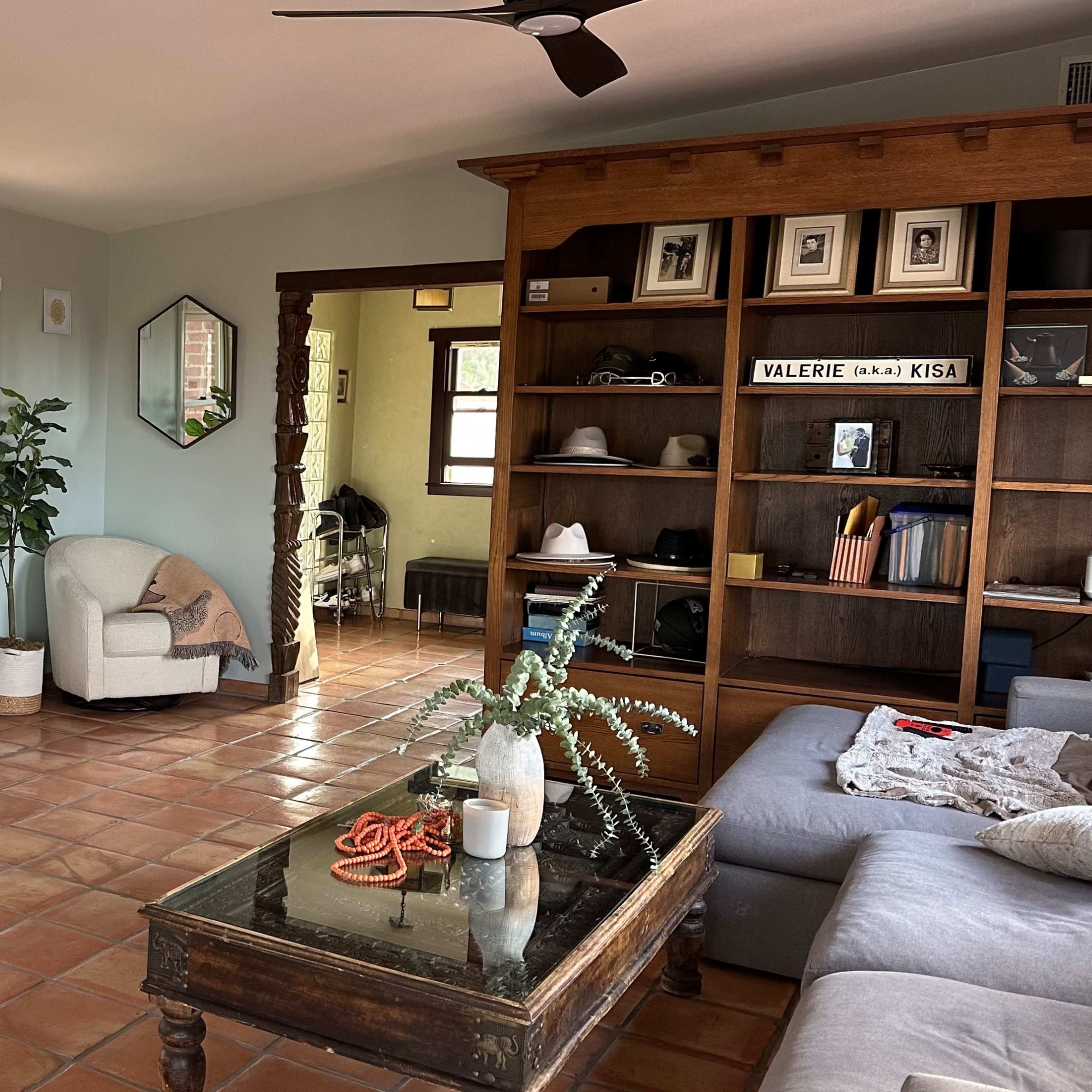 A spacious living room with a fireplace, a large TV mounted on the wall, a gray couch, and a wooden shelving unit filled with decorative items.