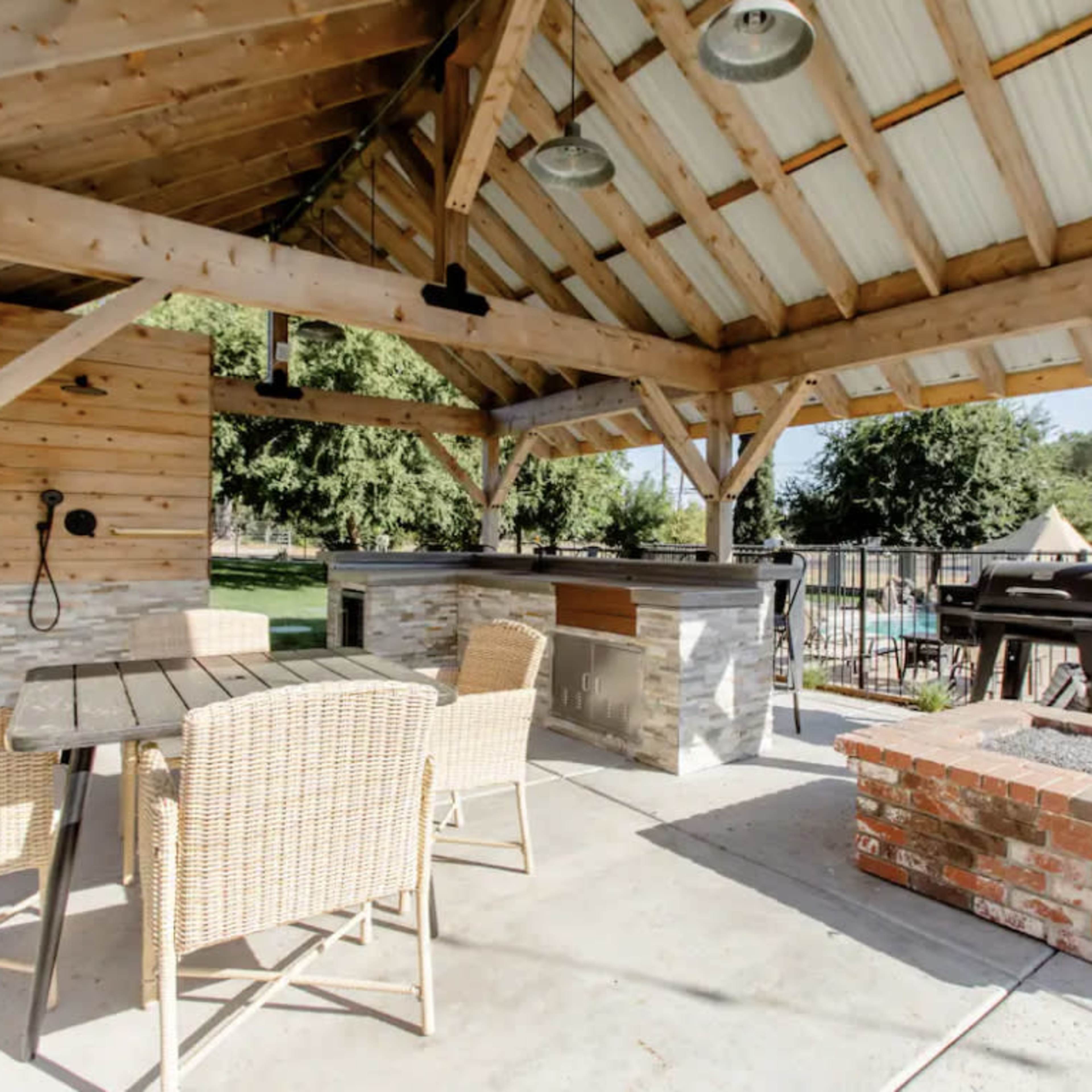 A shaded outdoor cooking area with a stone countertop, a grill, and seating arranged nearby.