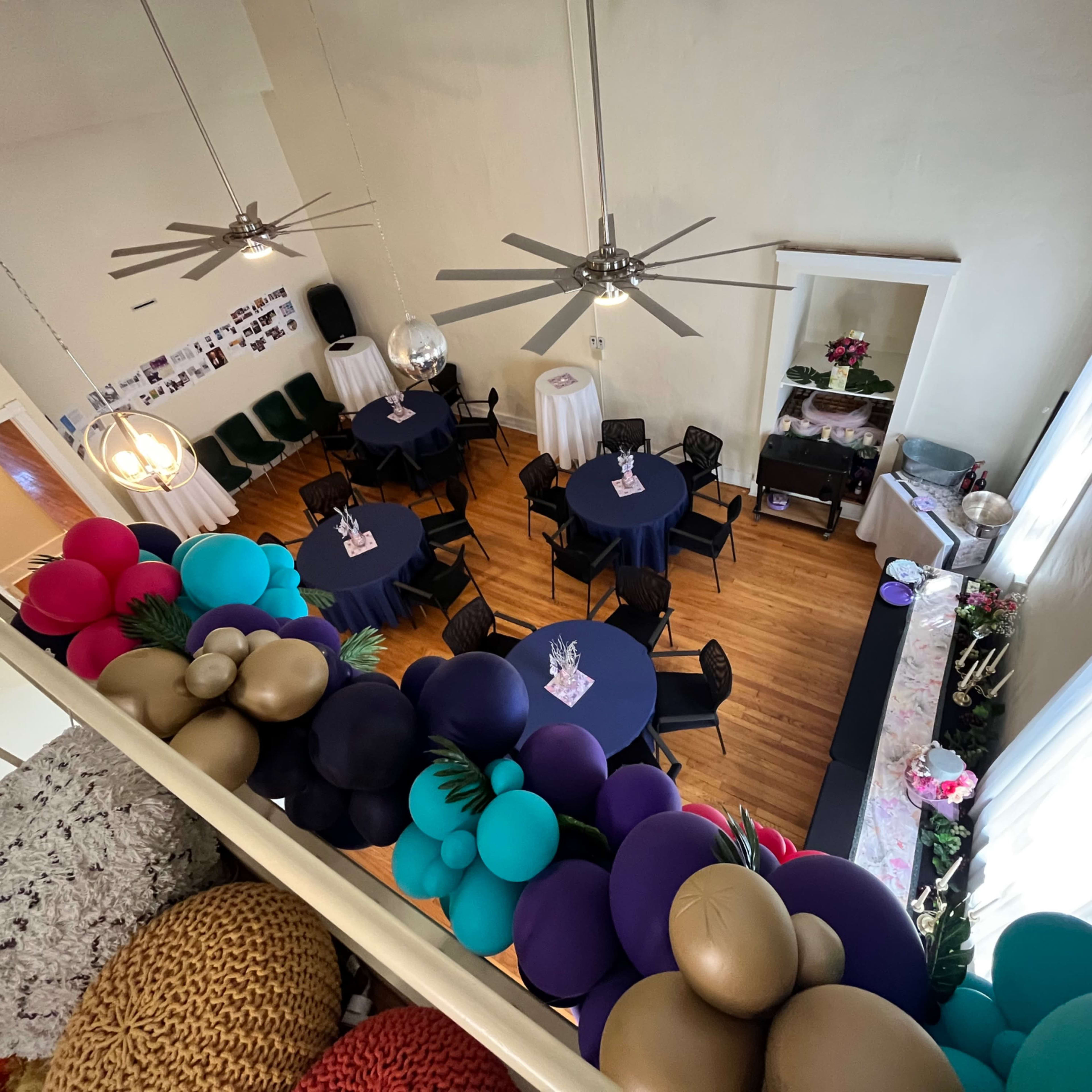 A decorated event space with round tables set for guests, large ceiling fans, and a colorful balloon garland along the balcony railing.