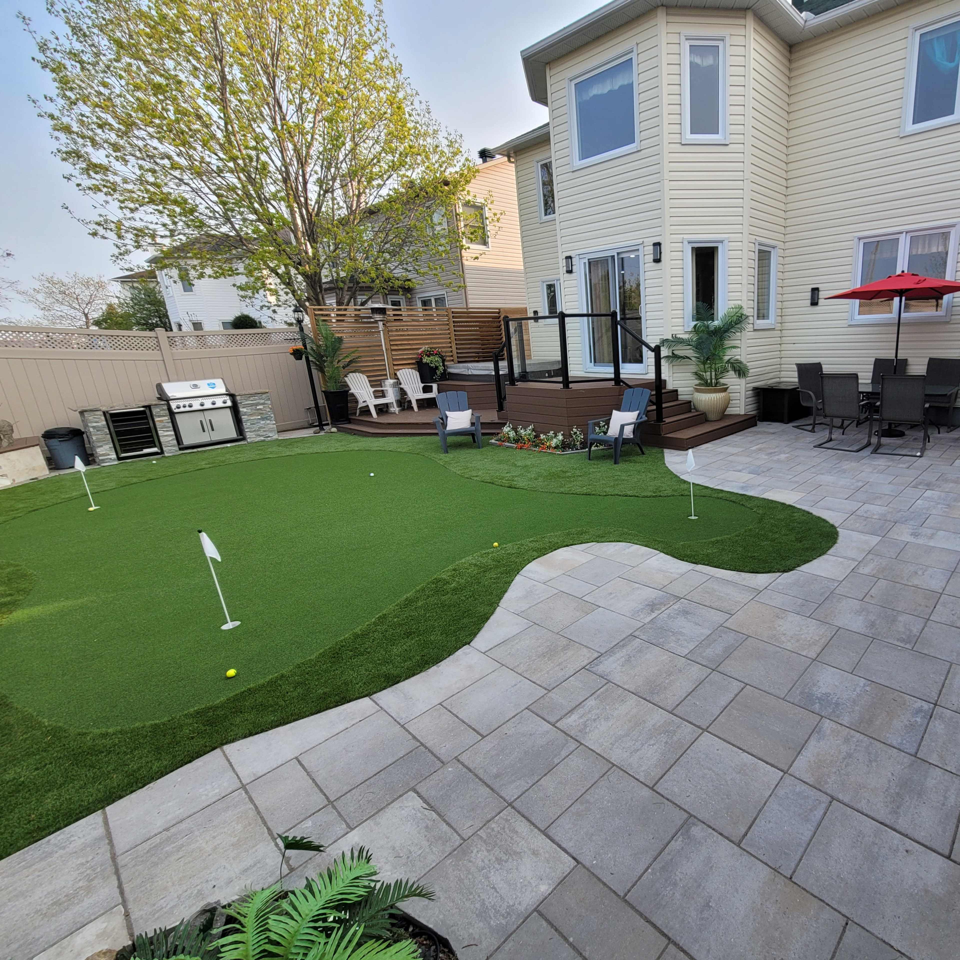 A landscaped backyard featuring a patio with seating, a putting green, and a house with a deck.