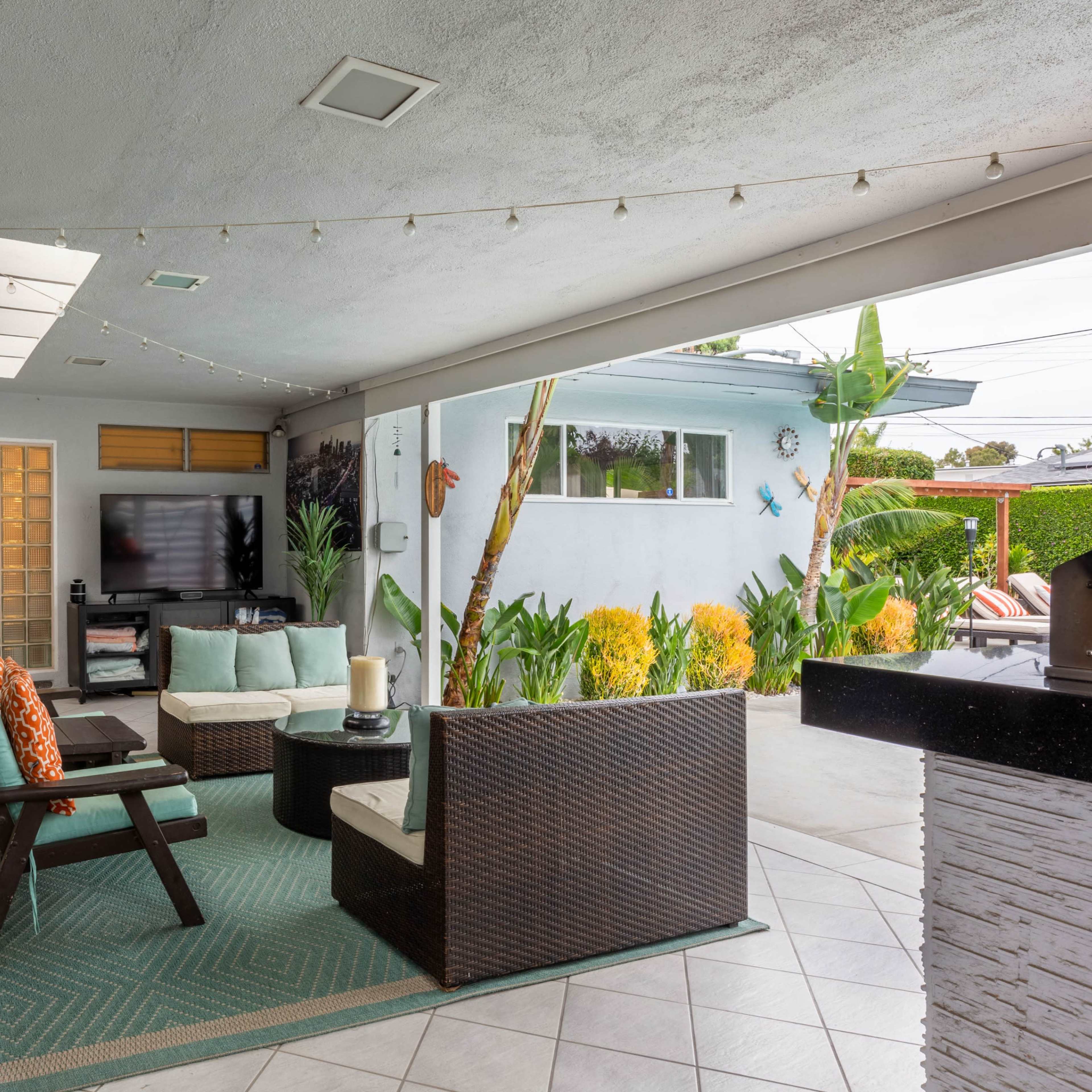A covered patio area with seating, a television, and decorative plants, leading to an outdoor space.