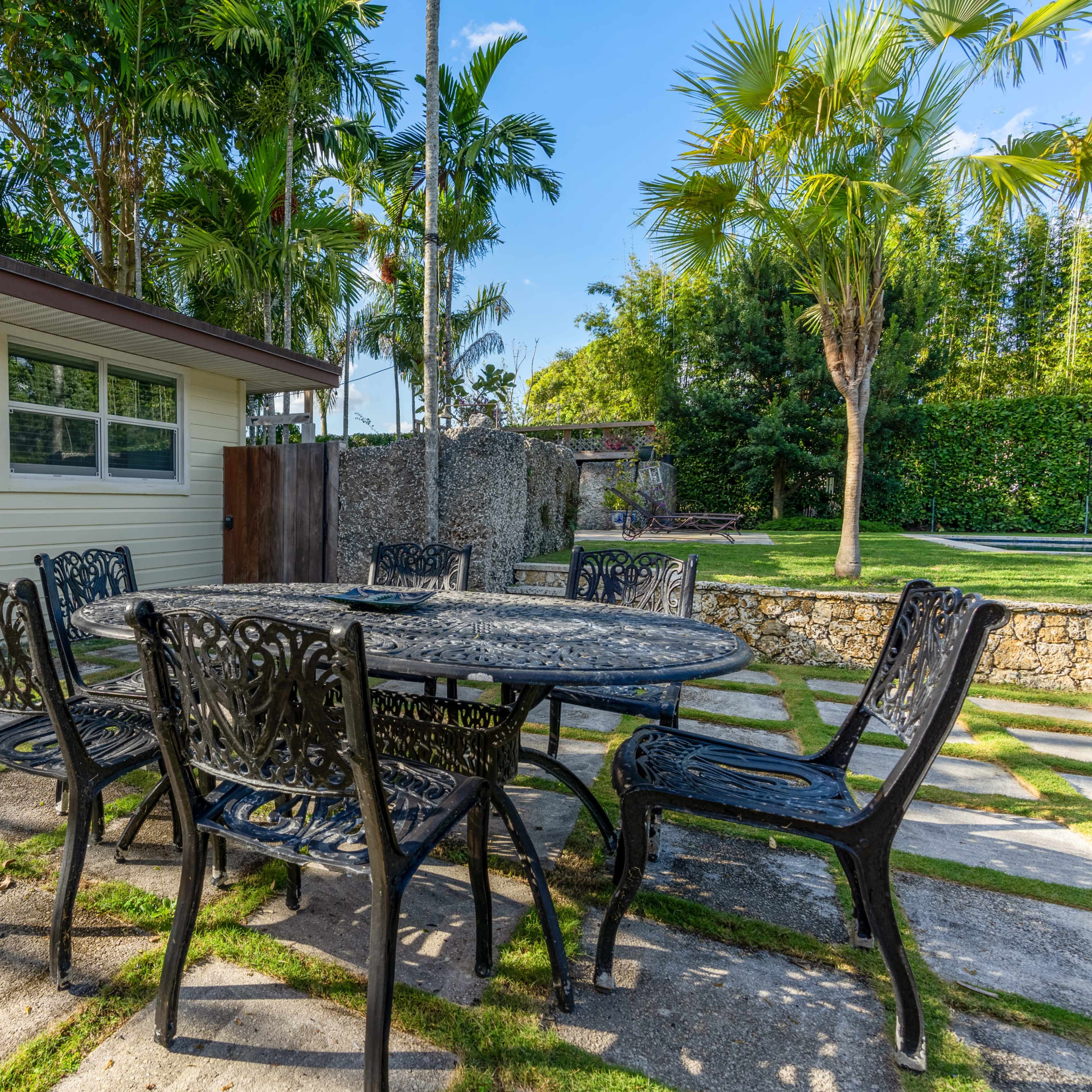 A circular metal dining table with six chairs is situated on a stone patio surrounded by palm trees and a modern shed.