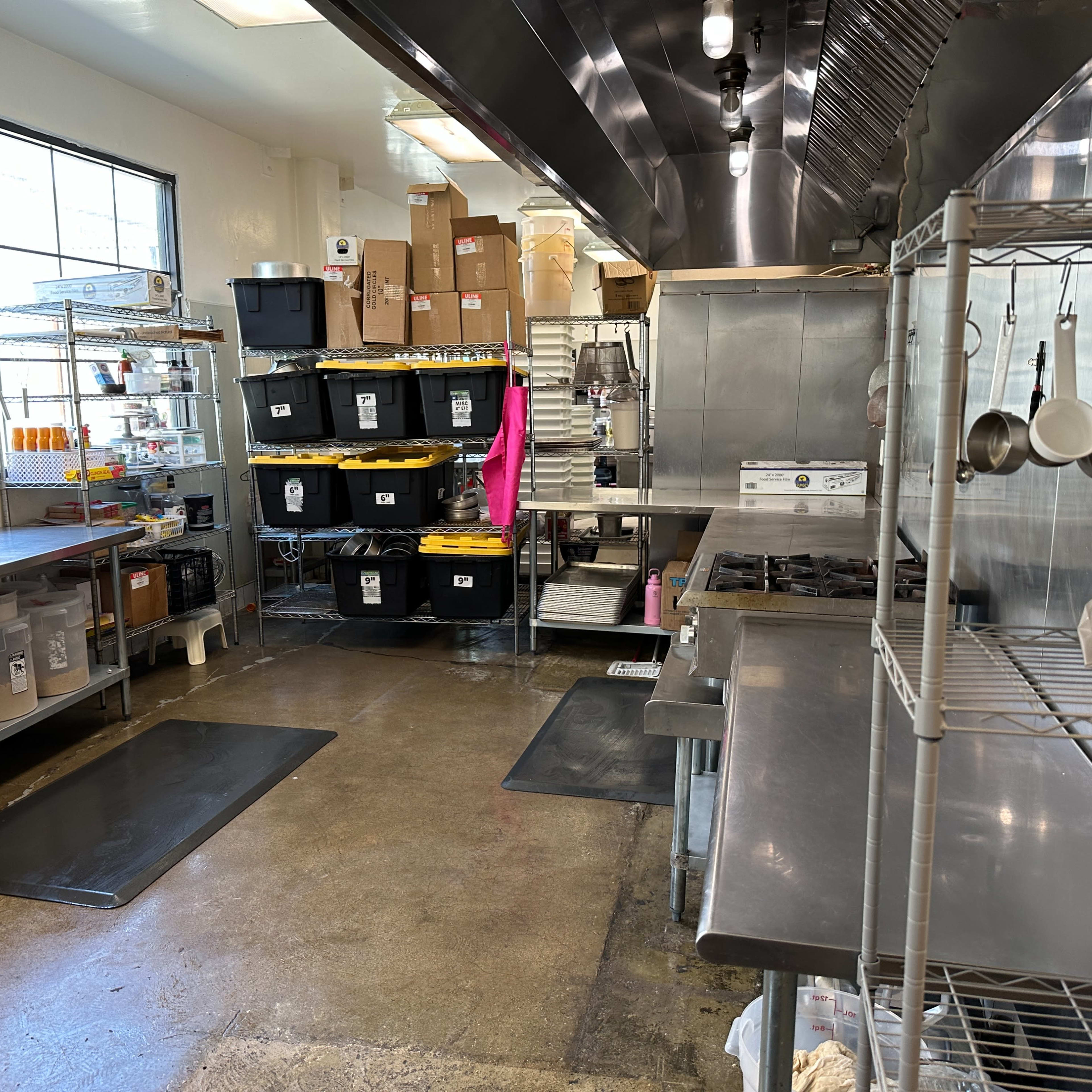 A commercial kitchen with stainless steel counters, shelves filled with storage bins, and various cooking equipment along the walls.