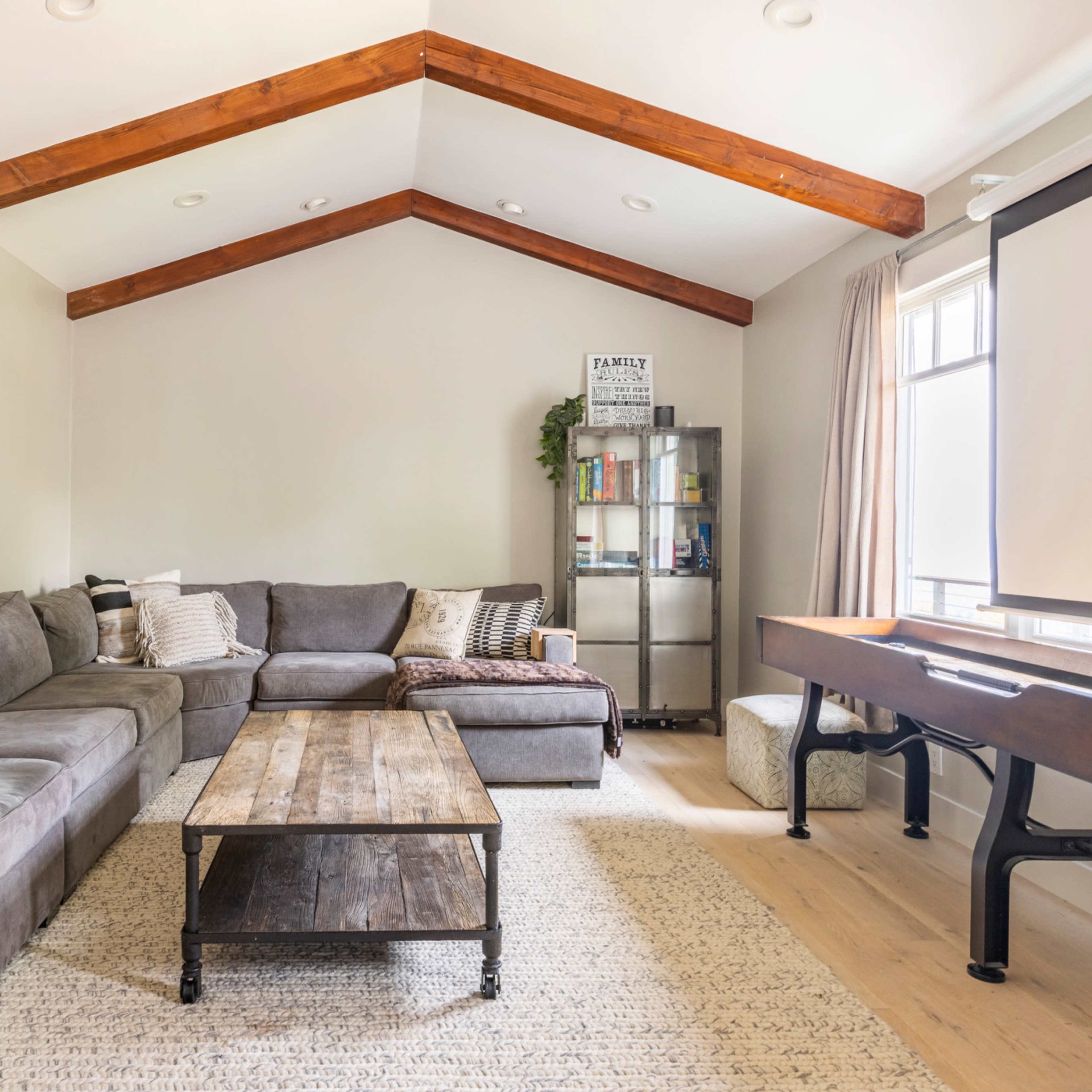 A spacious living room features a large sectional sofa, a wooden coffee table, a piano, and exposed wooden beams on the ceiling.