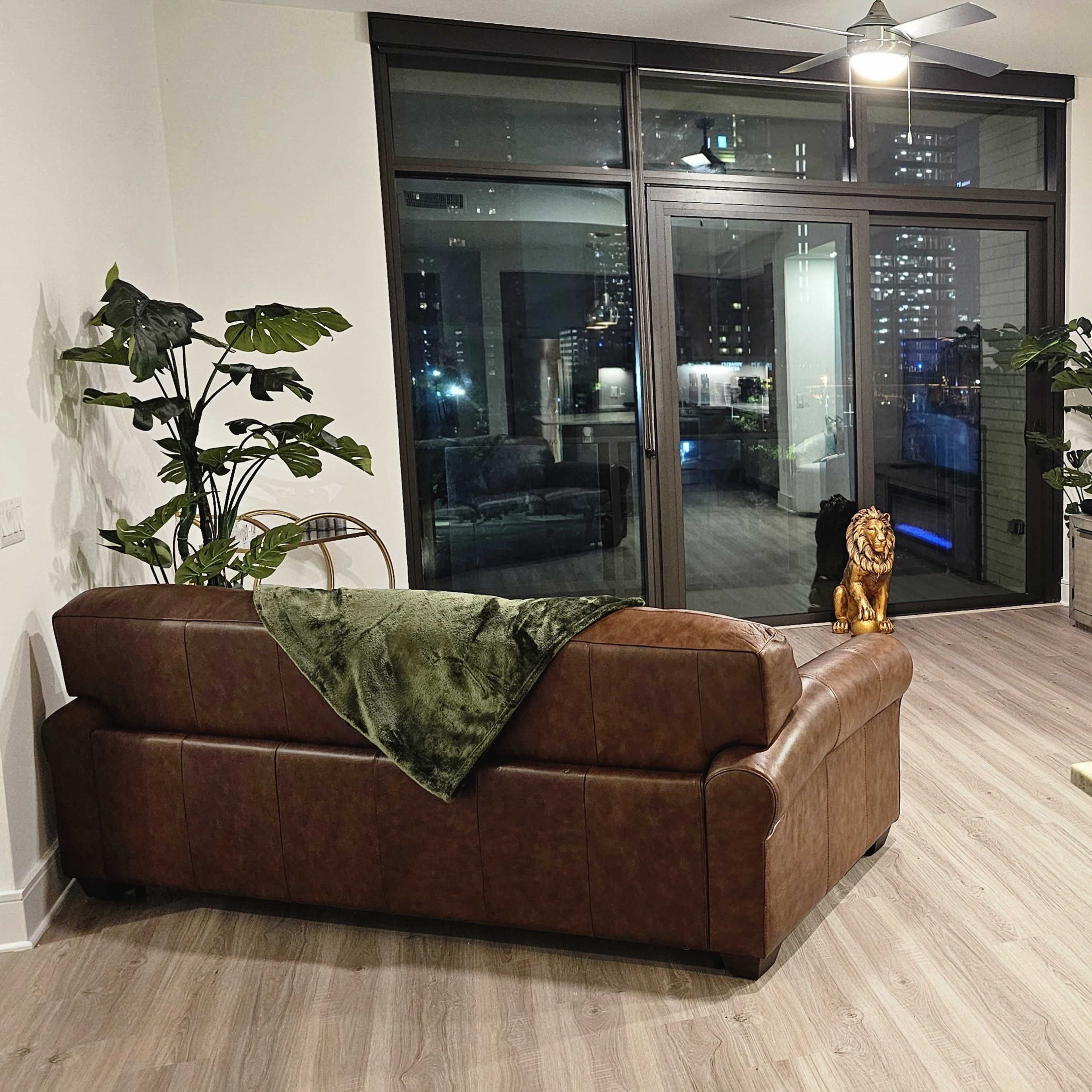 A modern living room features a brown leather sofa, a large television displaying a pool scene, and greenery on the walls, with a view of a city skyline through glass doors.