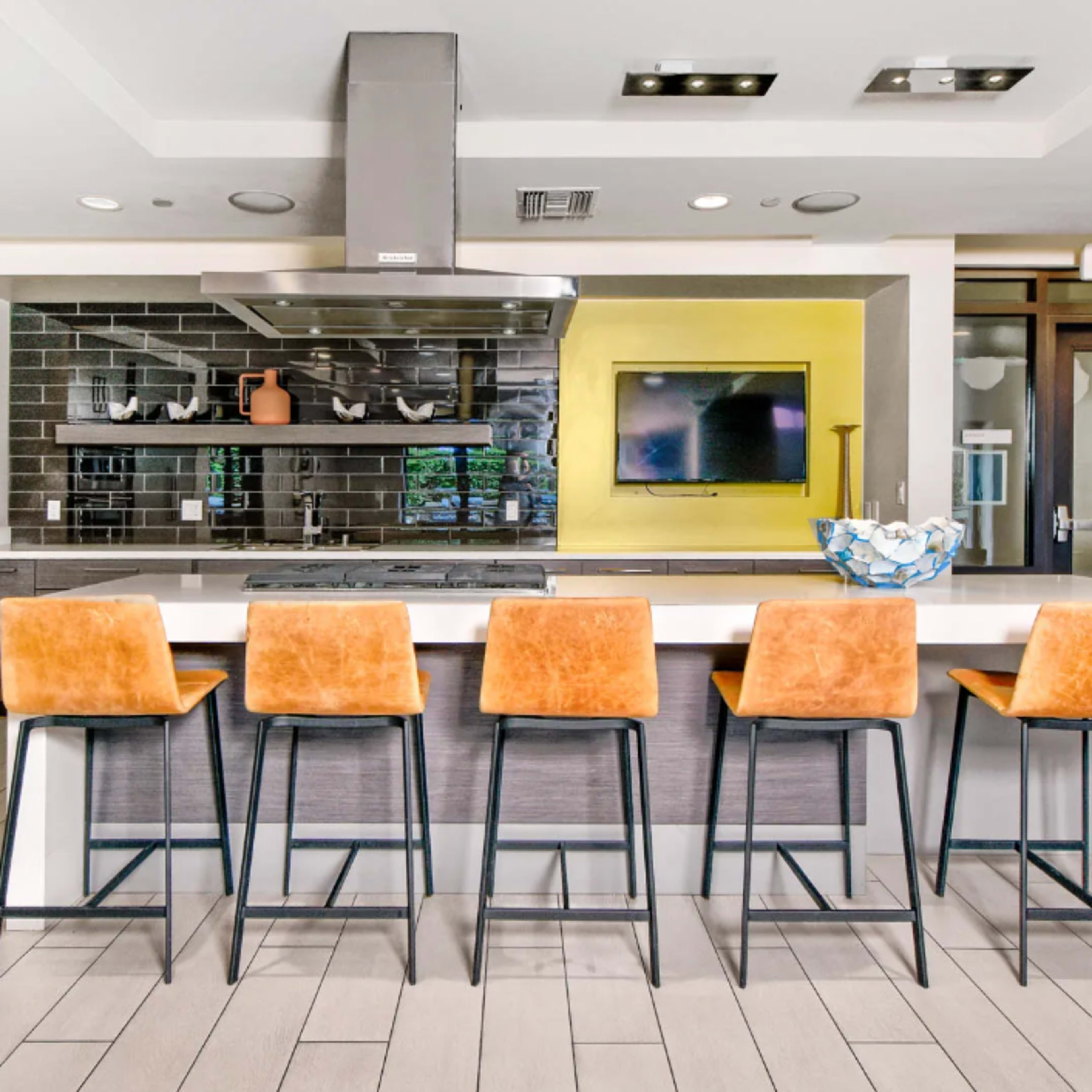 A modern kitchen features a large island with five orange bar stools and stainless steel appliances against a gray and black tiled backsplash.