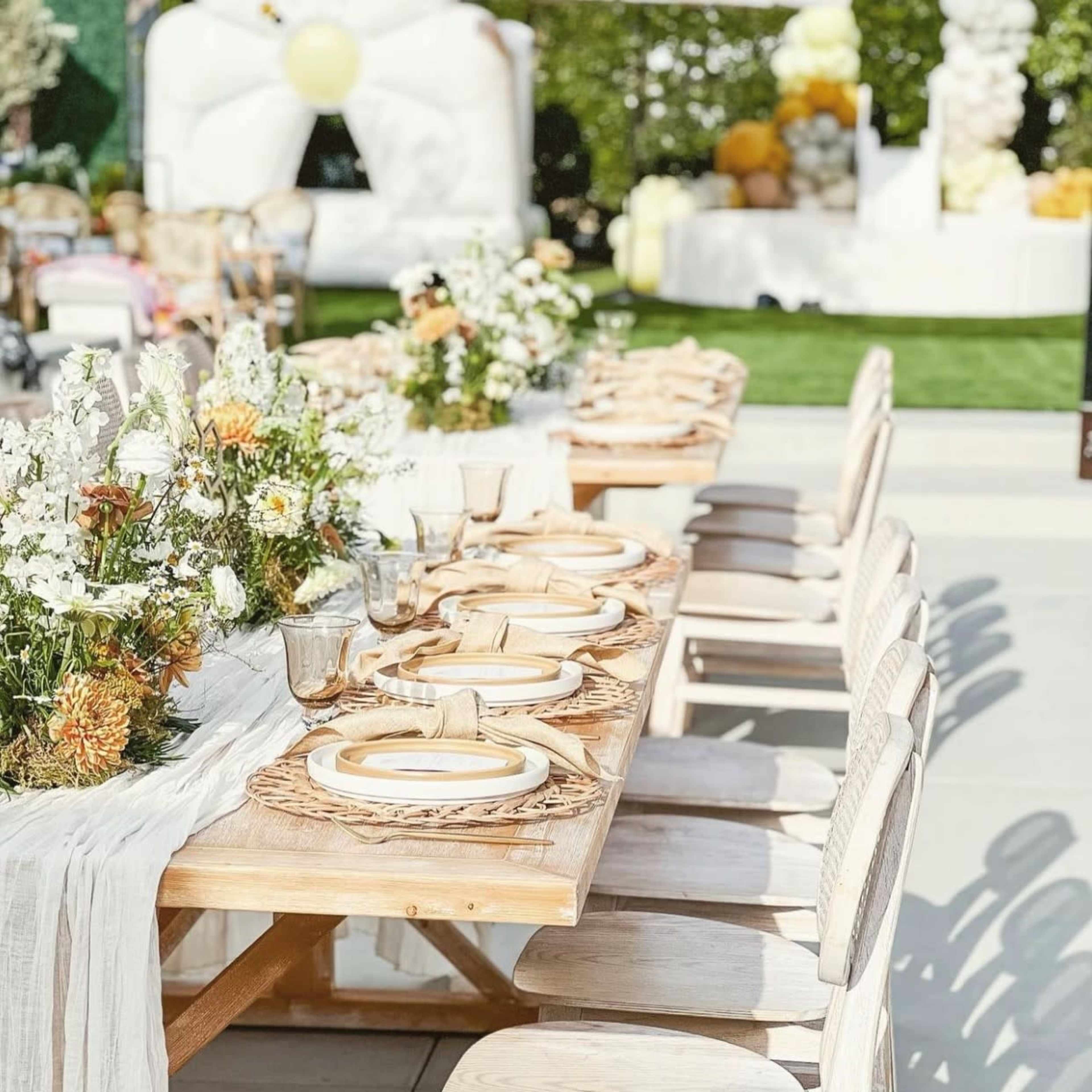 A long wooden table is set with plates and utensils, surrounded by empty chairs, and decorated with flowers in a manicured outdoor setting.