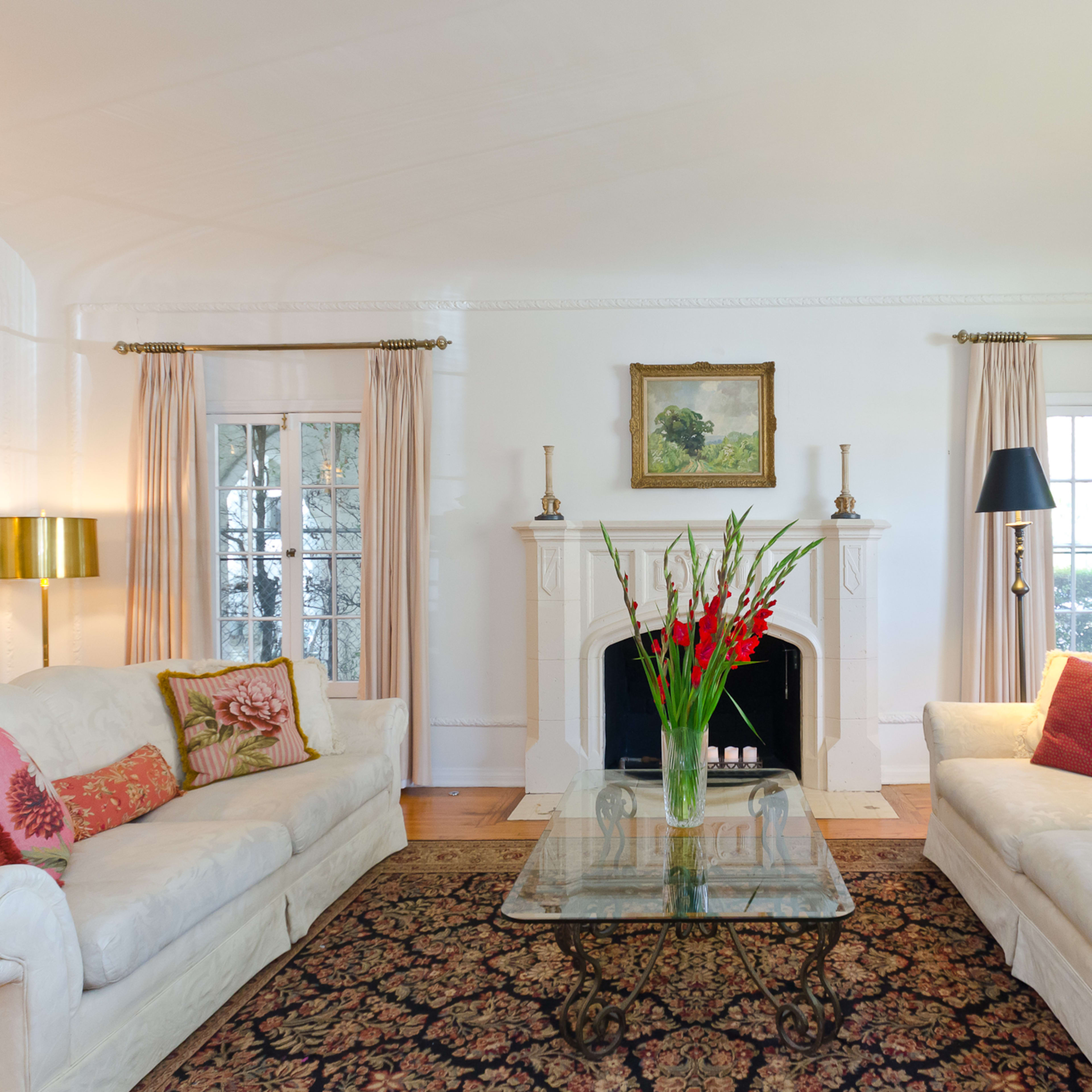 A bright living room features two white sofas with decorative pillows, a glass coffee table, and a fireplace, framed by large windows and elegant curtains.