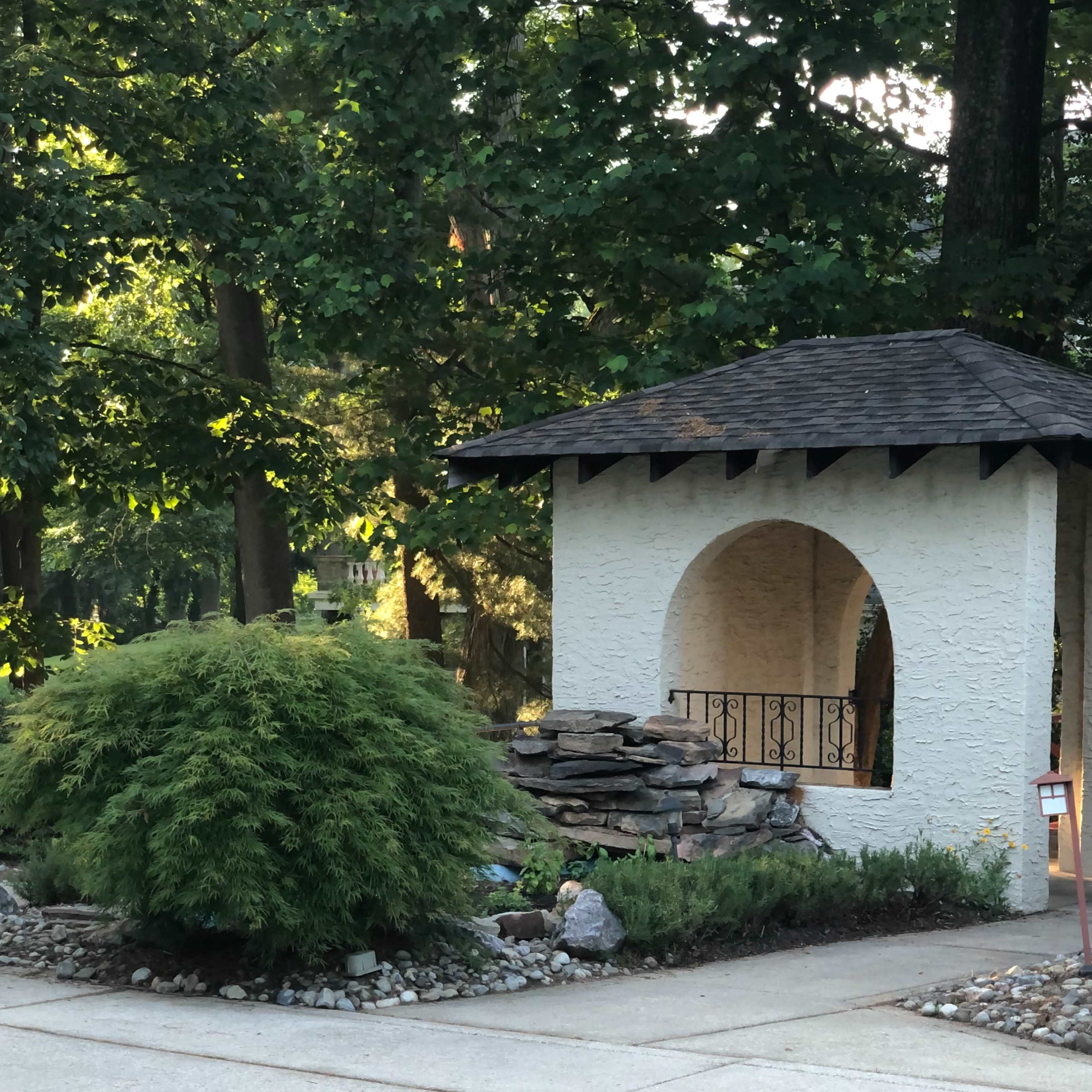 A white gazebo with a sloped roof sits beside a pool, surrounded by greenery and stone landscaping.