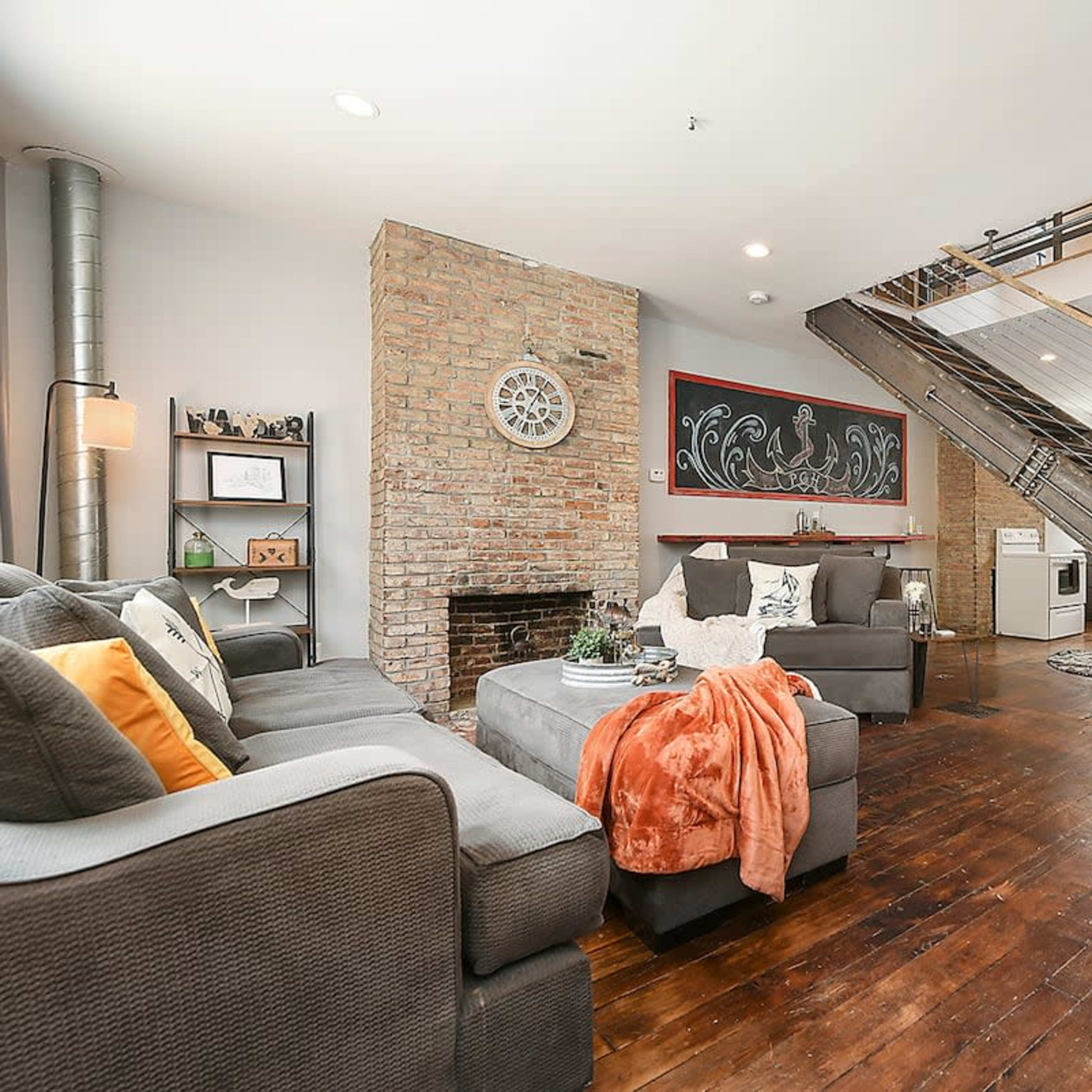 A modern living room with a brick wall, a gray couch, a fireplace, and a staircase leading to an upper level.