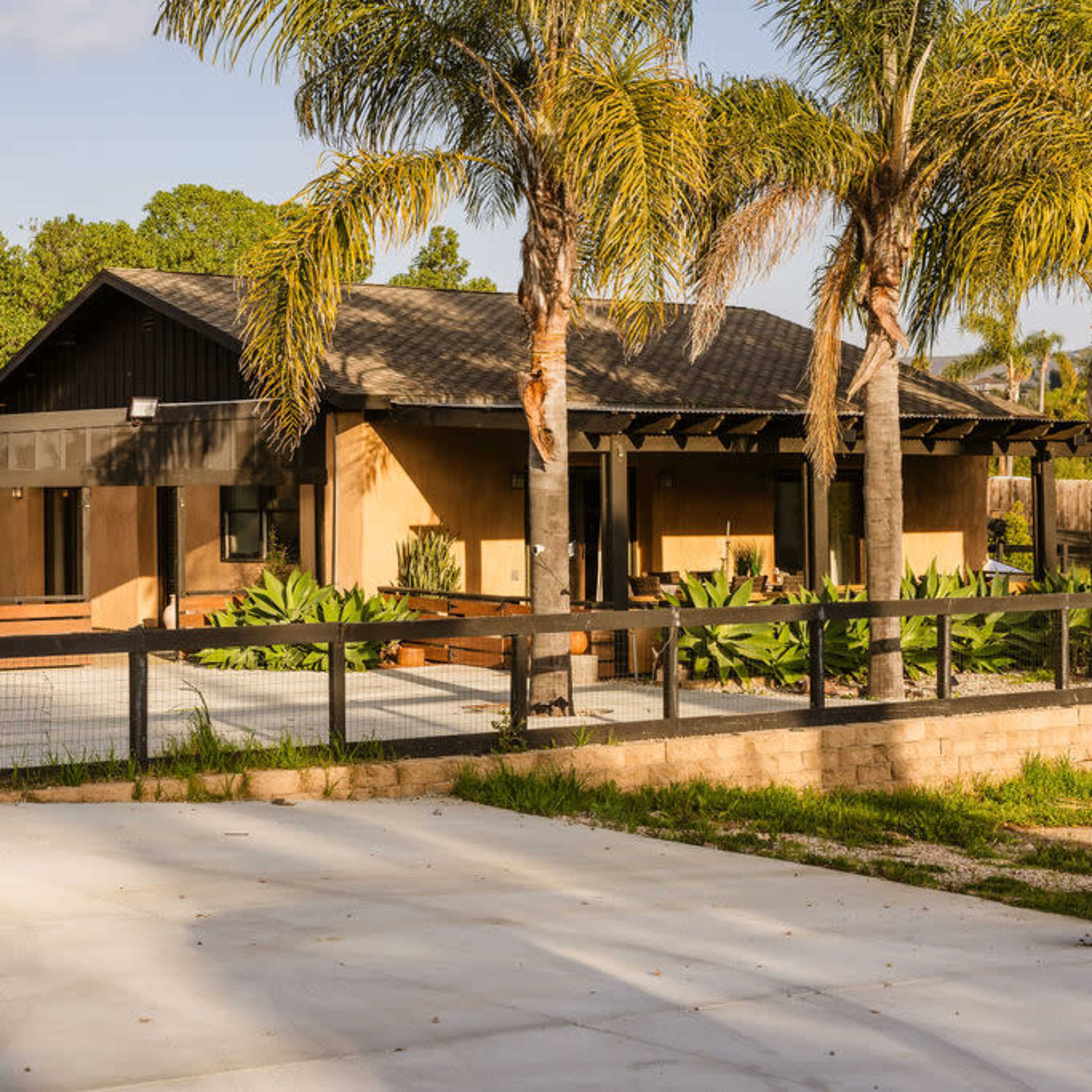 A modern single-story house with a wooden deck and palm trees is surrounded by greenery and a fenced patio area.