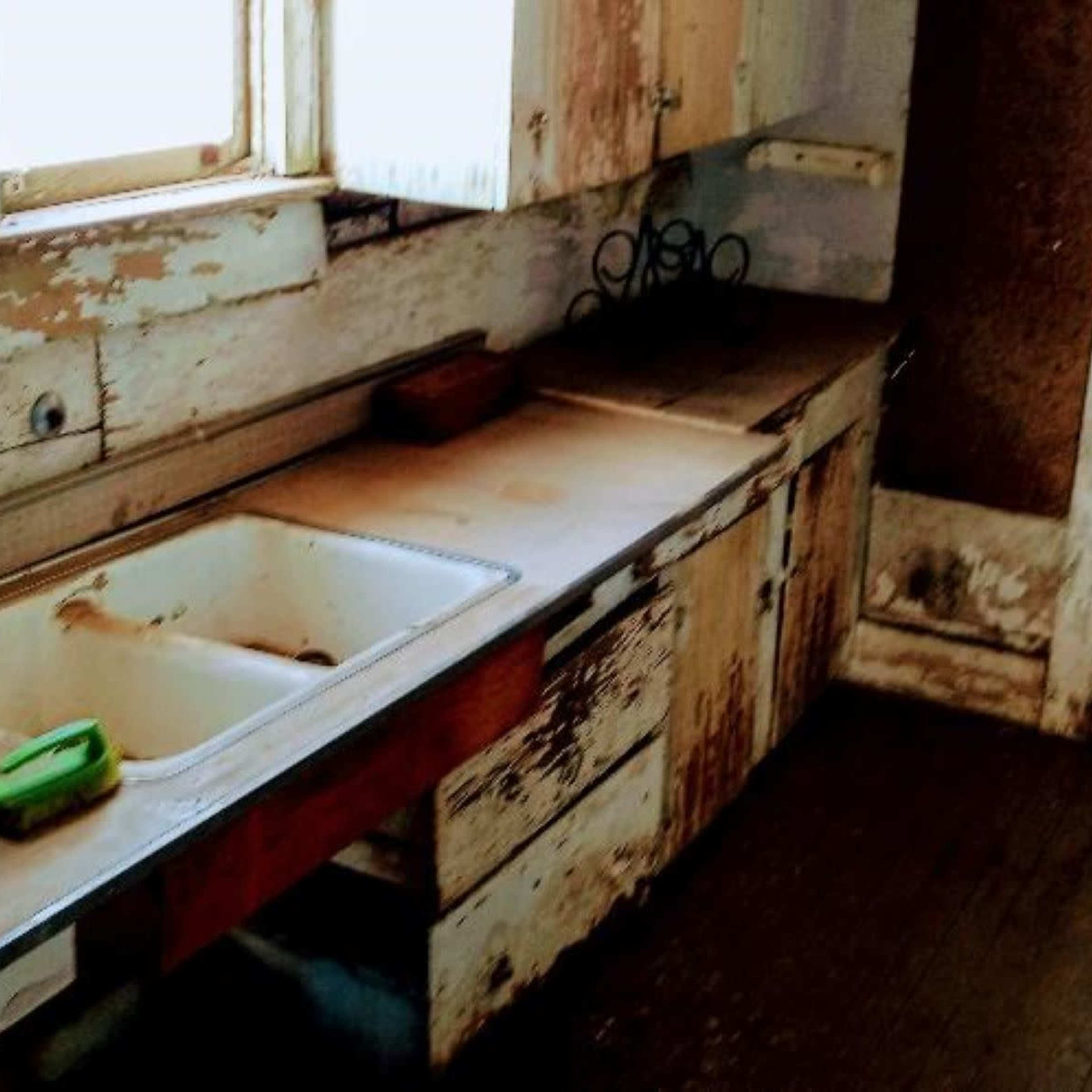A worn kitchen with peeling paint on the cabinets and an empty sink beside a microwave and a door.
