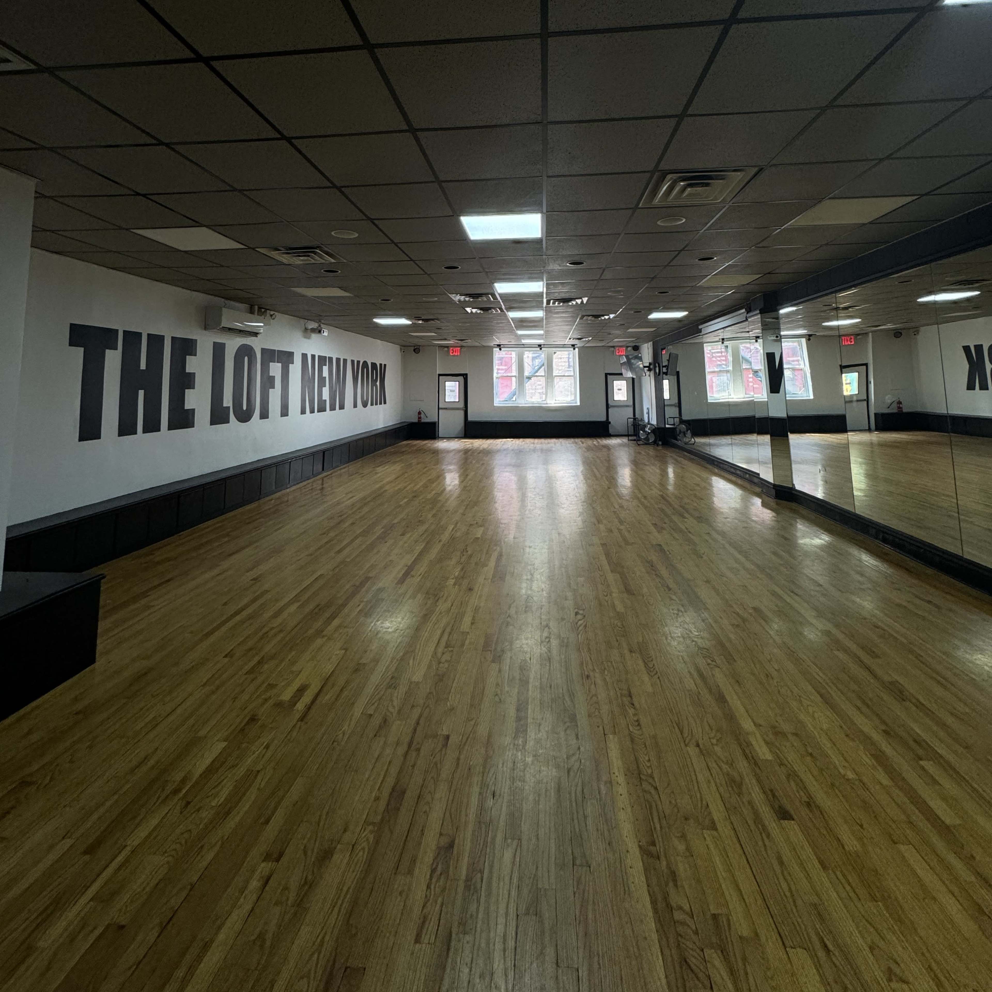 An empty dance studio with wooden flooring and a large mirror on one wall, featuring the words "THE LOFT NEW YORK" on the opposite wall.
