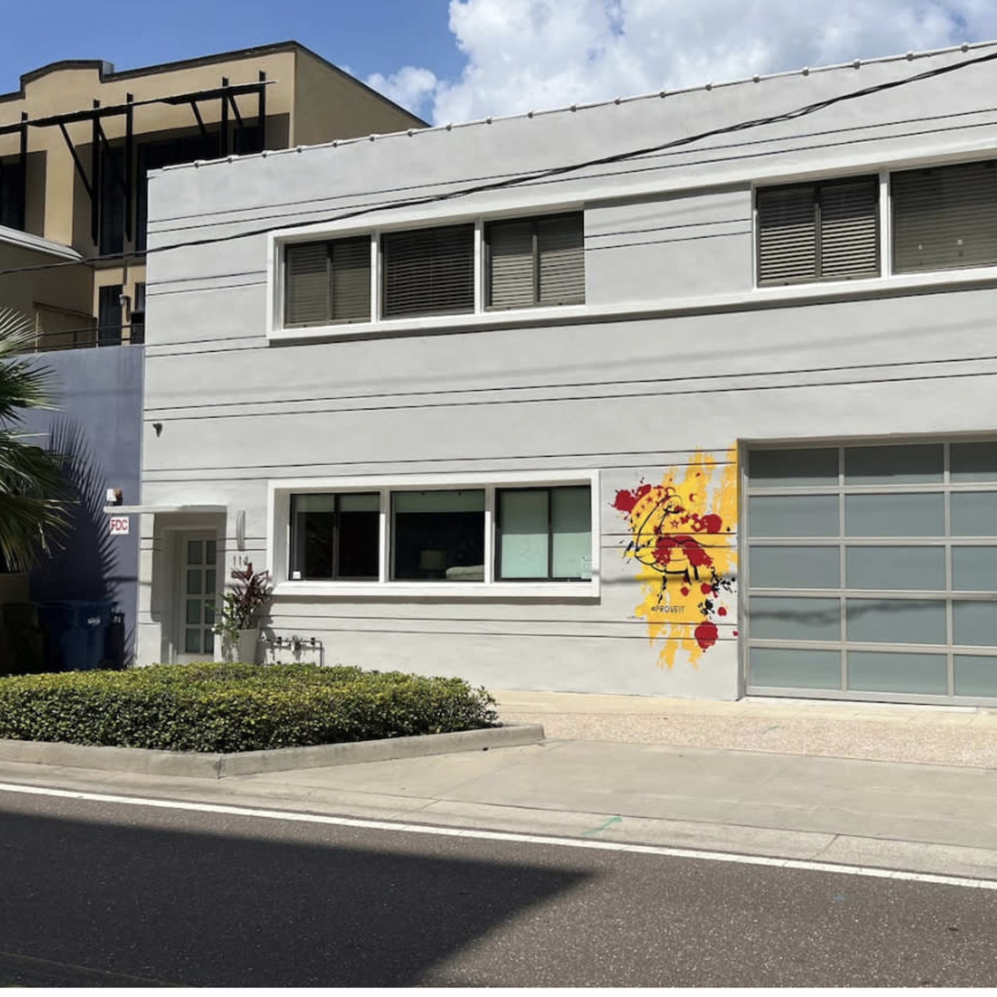 A modern gray building with large windows and a colorful mural on one side, located on a clean street with parked bins.