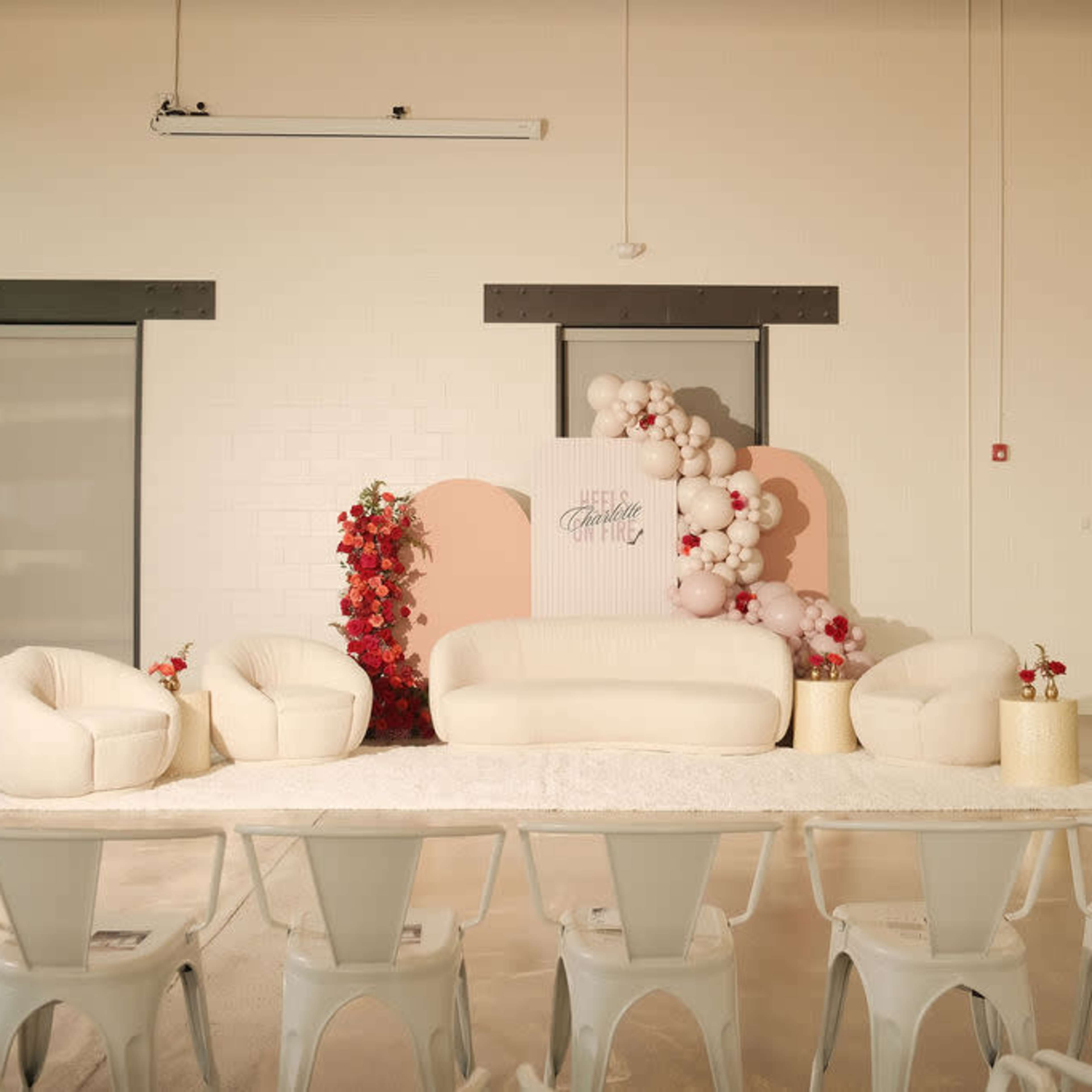 A staged event space with a white sofa, additional seating, a backdrop featuring balloons and floral arrangements, and rows of chairs facing the setup.