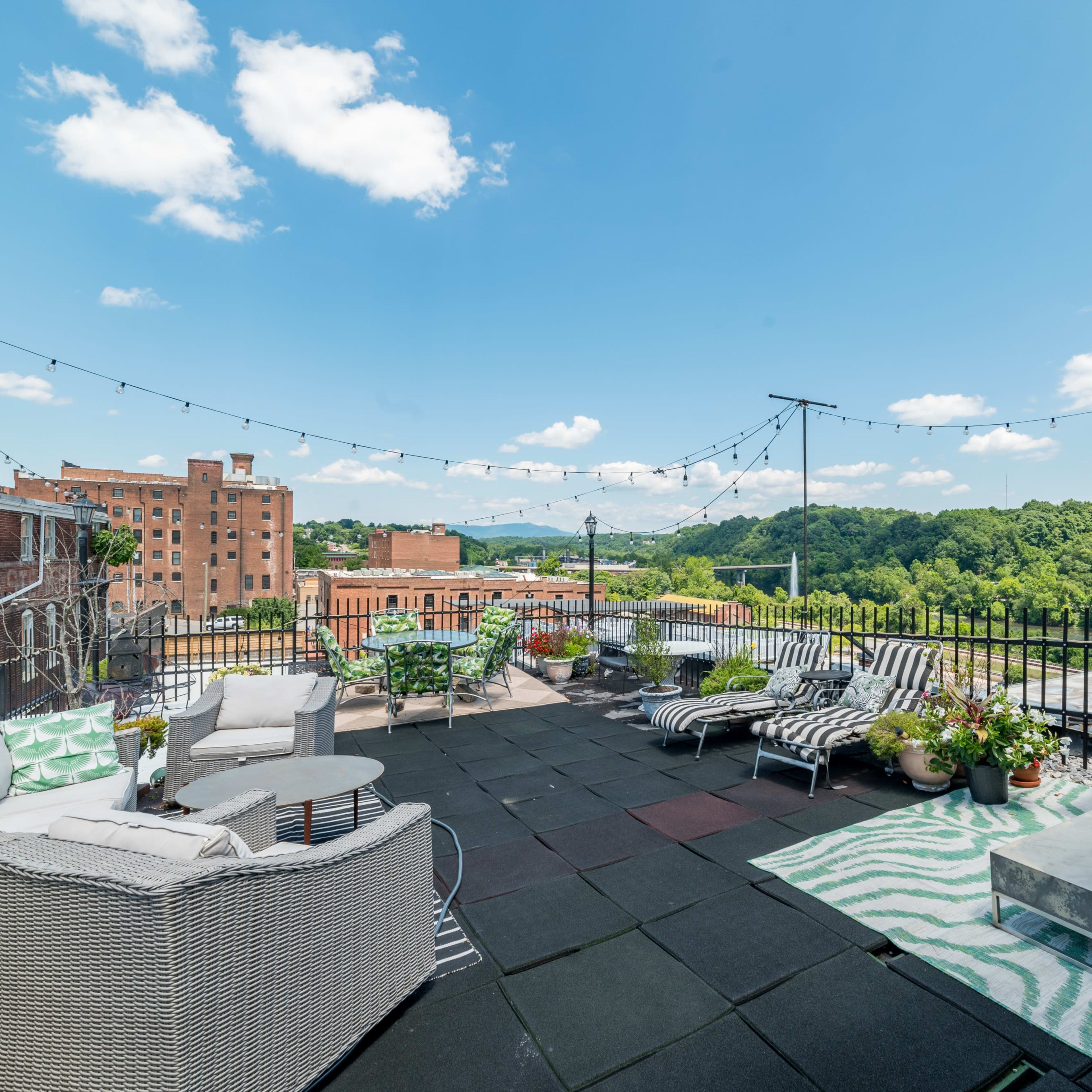 A spacious rooftop terrace features seating areas, decorative plants, and a view of distant hills under a clear sky with scattered clouds.
