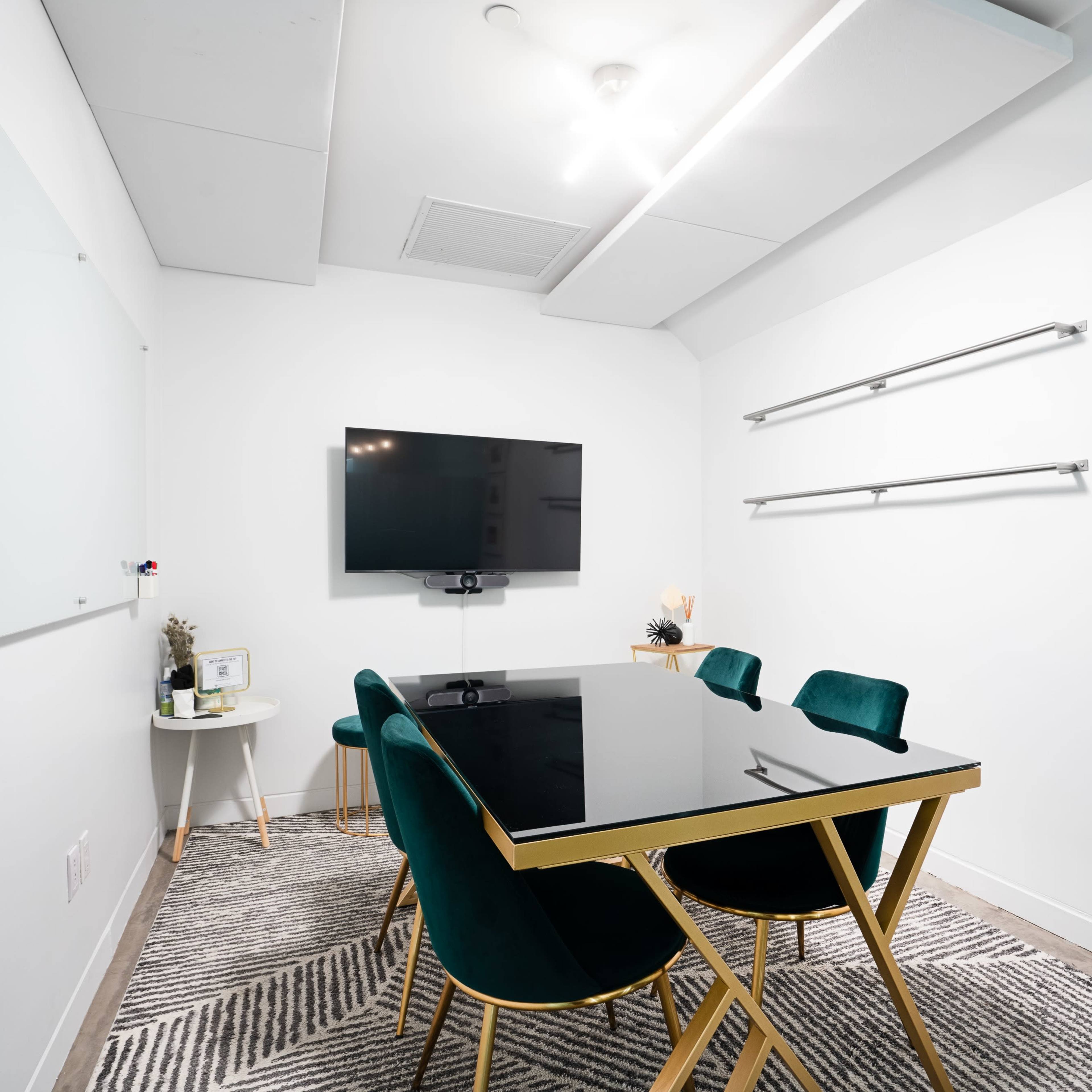 A modern conference room features a black glass table surrounded by green chairs, a television mounted on the wall, and minimalistic decor.