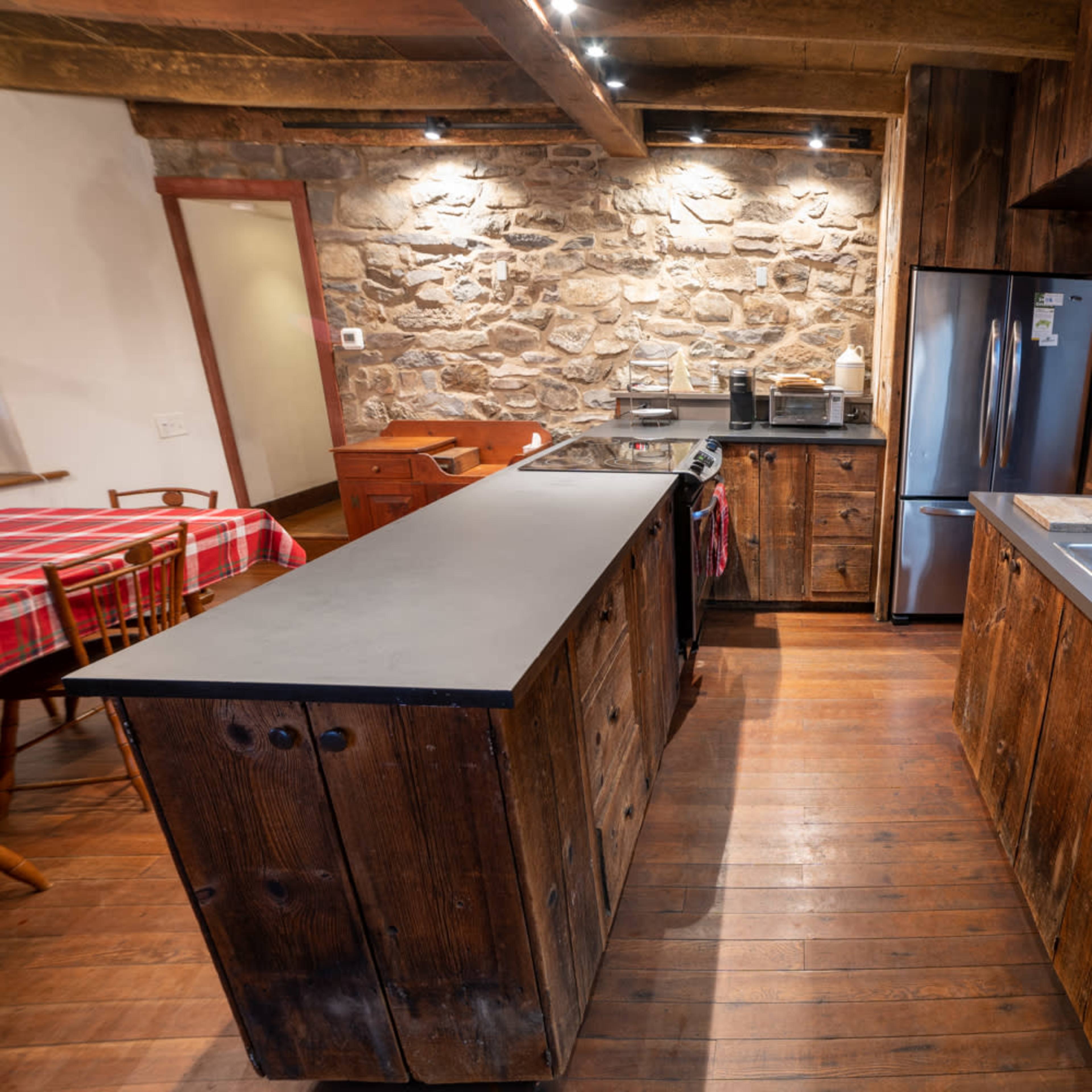 A rustic kitchen with wooden cabinetry, a stone wall, stainless steel appliances, and a central island with a black countertop.