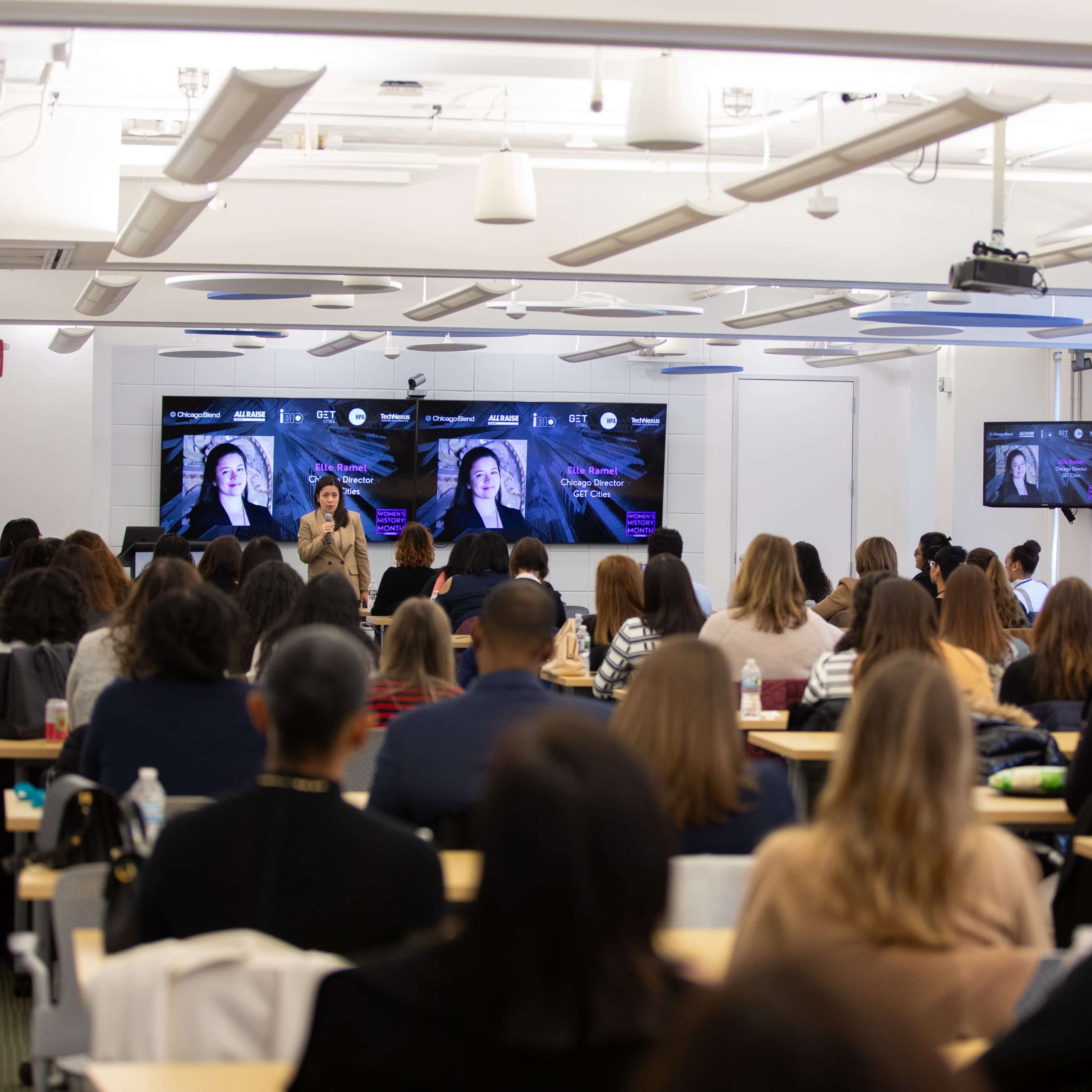 A group of people sits in a classroom setting, facing a presenter and screens displaying visuals.