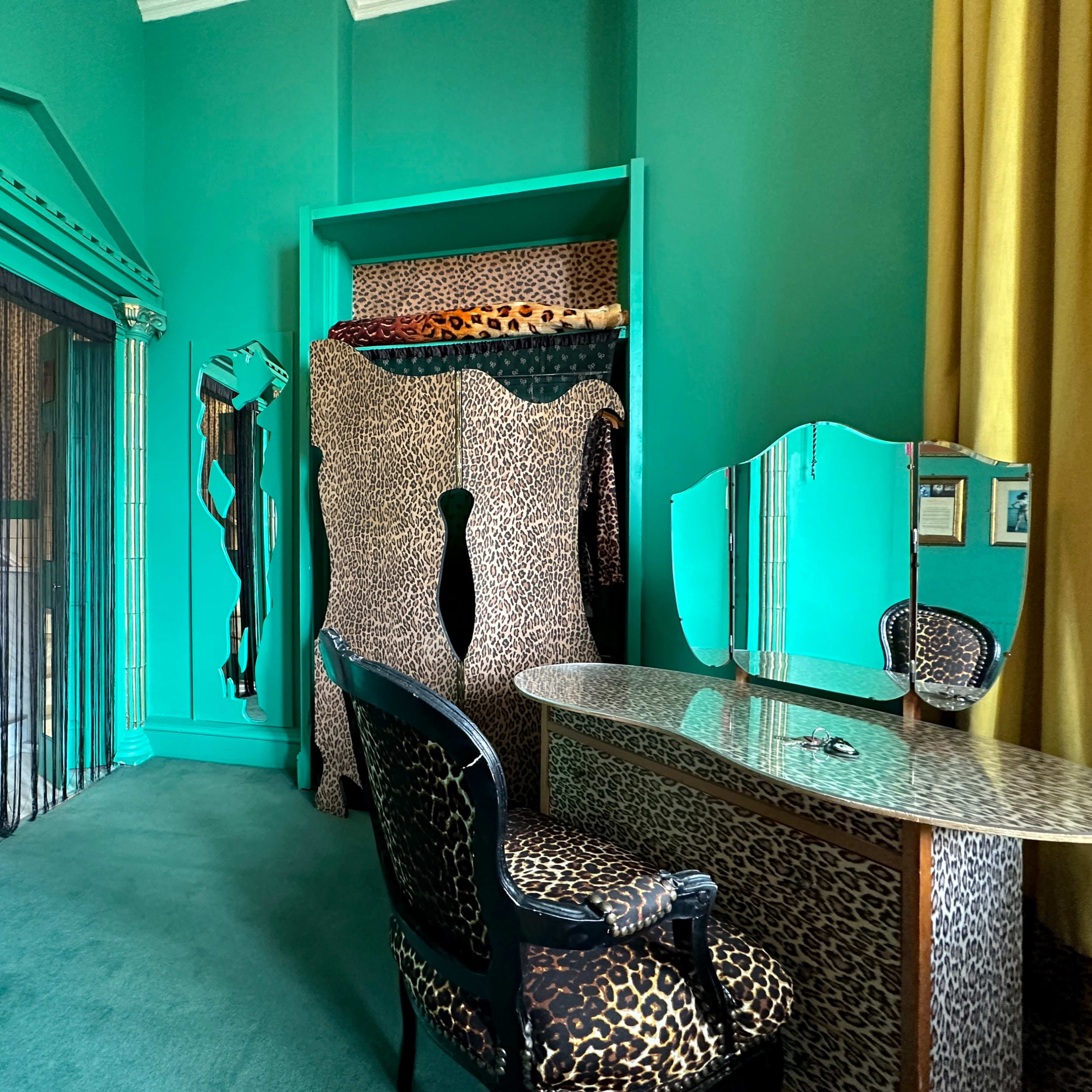 The room features green walls and leopard-print furniture, including a chair and a vanity, with a decorative mirror and a closet area.