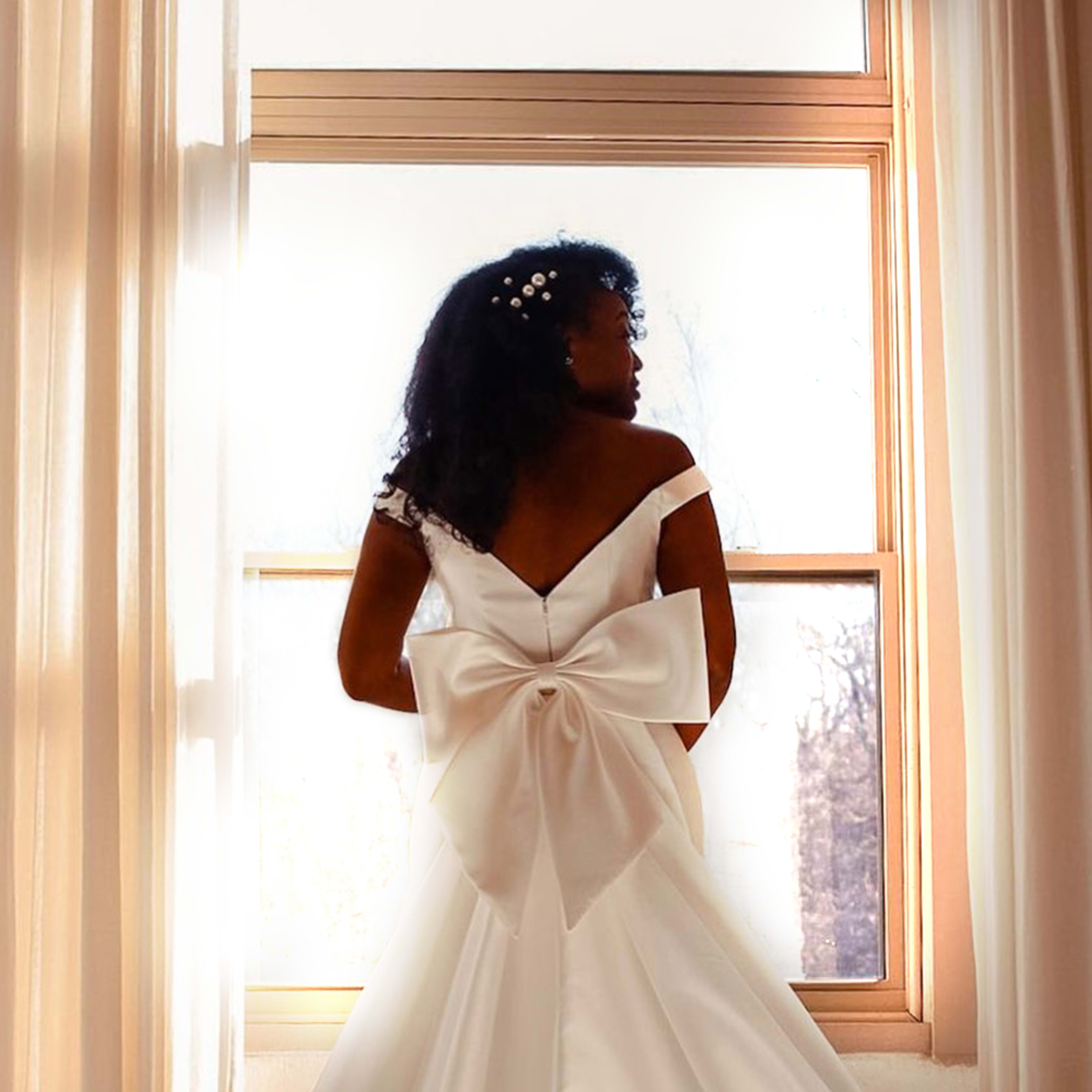 A woman in a flowing white wedding dress with a large bow stands by a window, facing away from the camera.