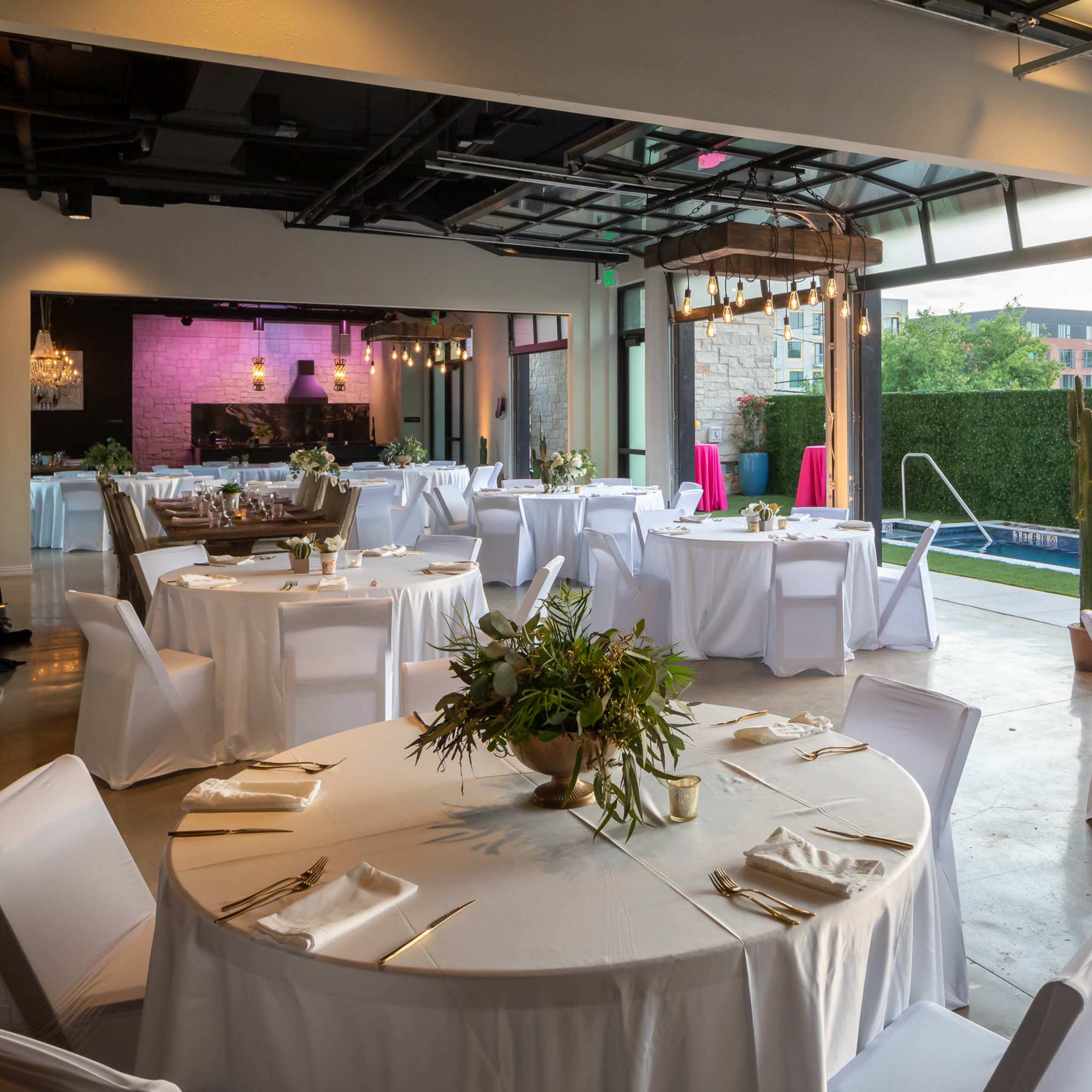 A modern event space with round tables set for dining, surrounded by large windows open to a pool area outside.