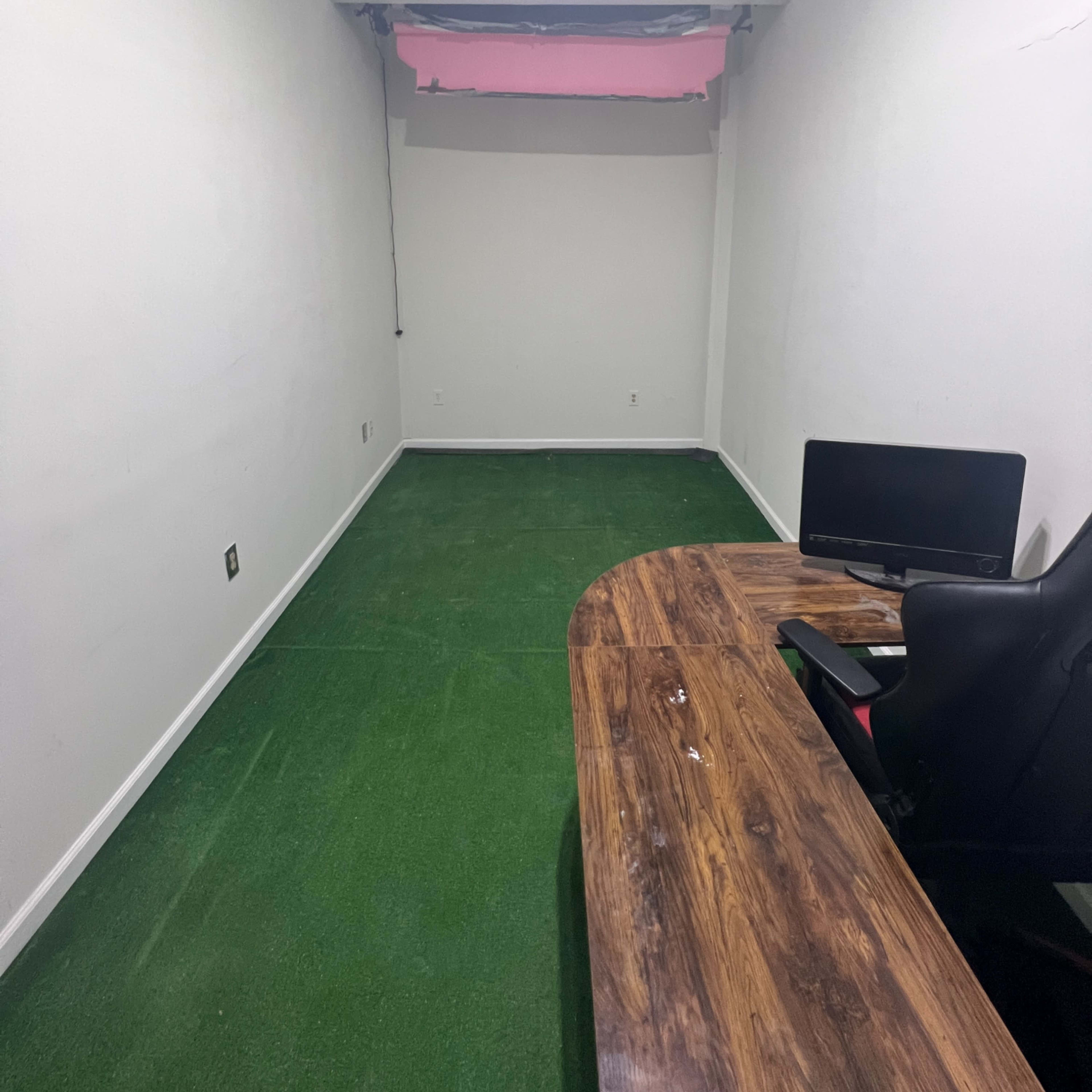 A small, empty room with green carpet, a wooden desk in the corner, a black chair, and a computer monitor.