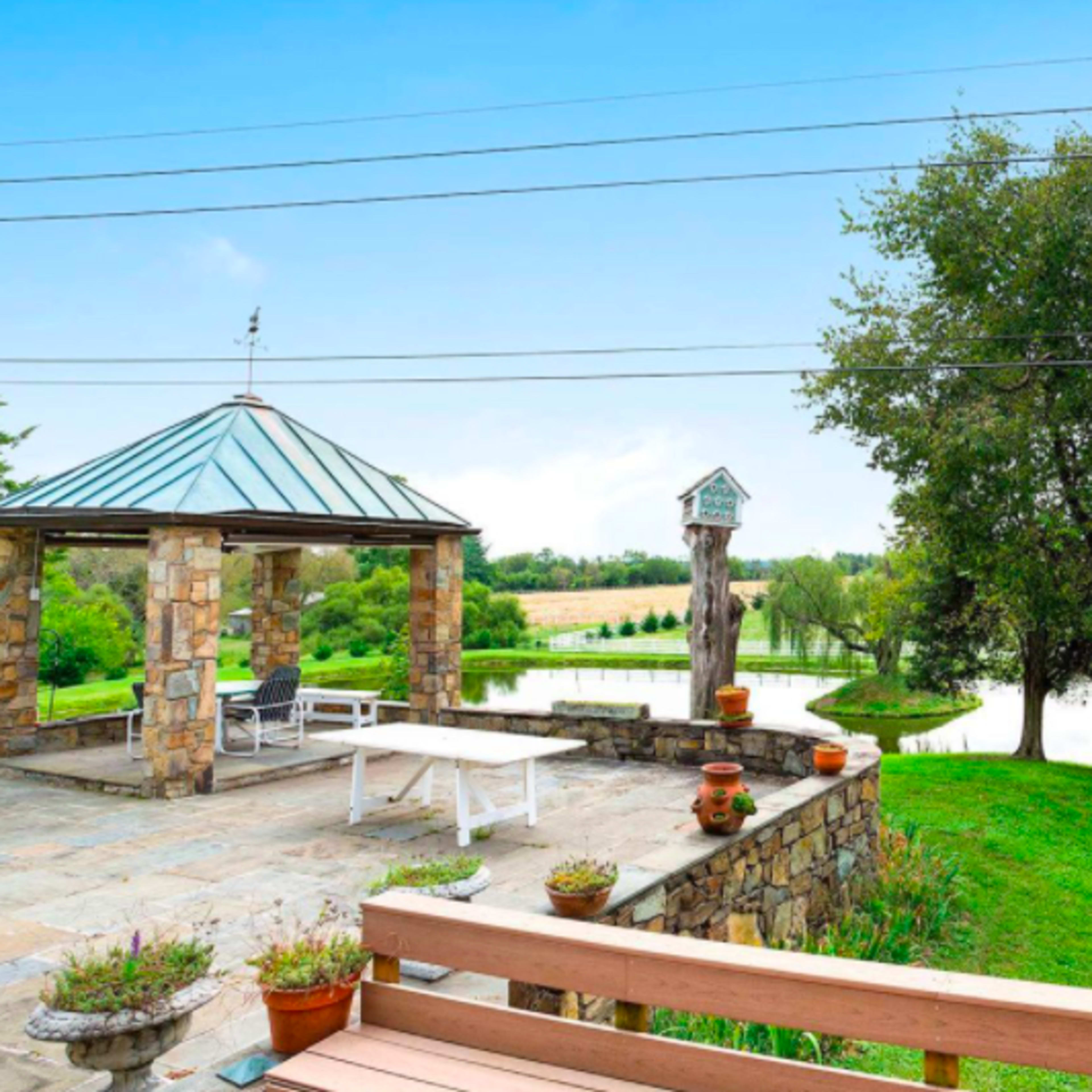A stone gazebo overlooking a pond, surrounded by green grass and trees, with a table and chairs on a patio area.
